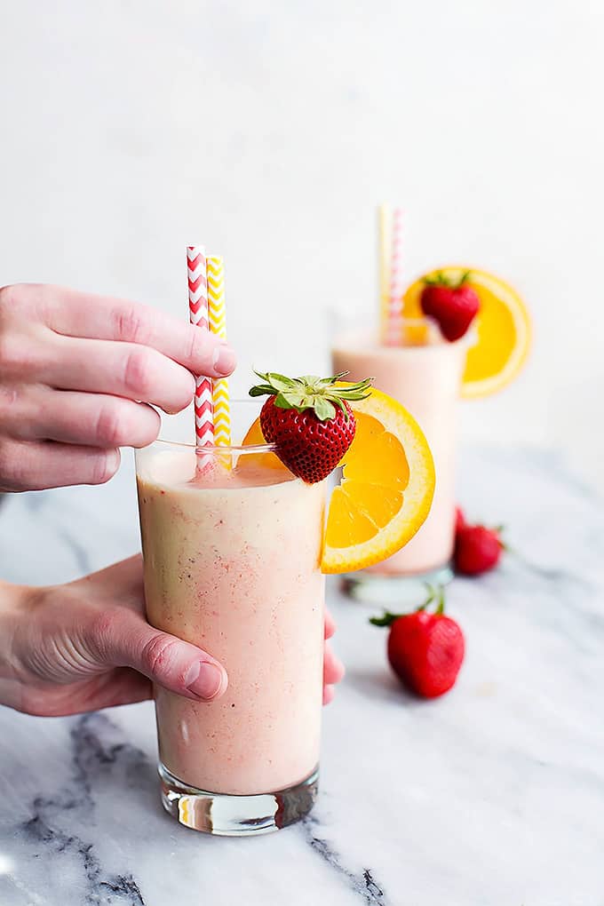 hands grabbing a glass of strawberry orange Julius  and the two straws on top with another glass in the background.