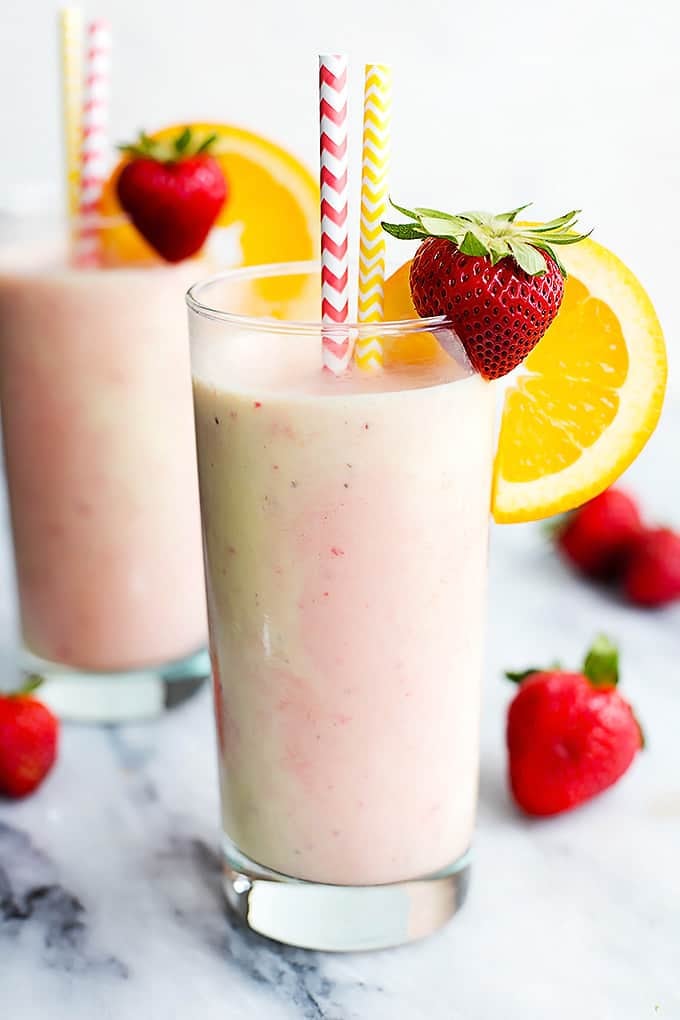 a glass of strawberry orange Julius with two straws and a strawberry and orange wheel on the side of the glass with another glass in the background both surrounded by strawberries.