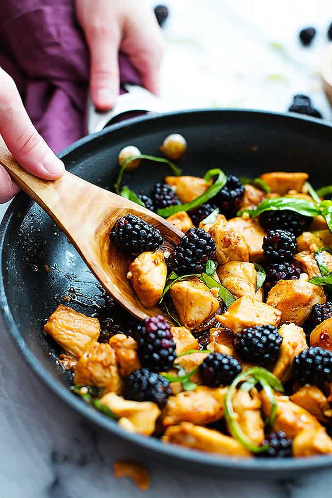 a hand scooping some Thai blackberry basil chicken out of a skillet with a wooden serving spoon.