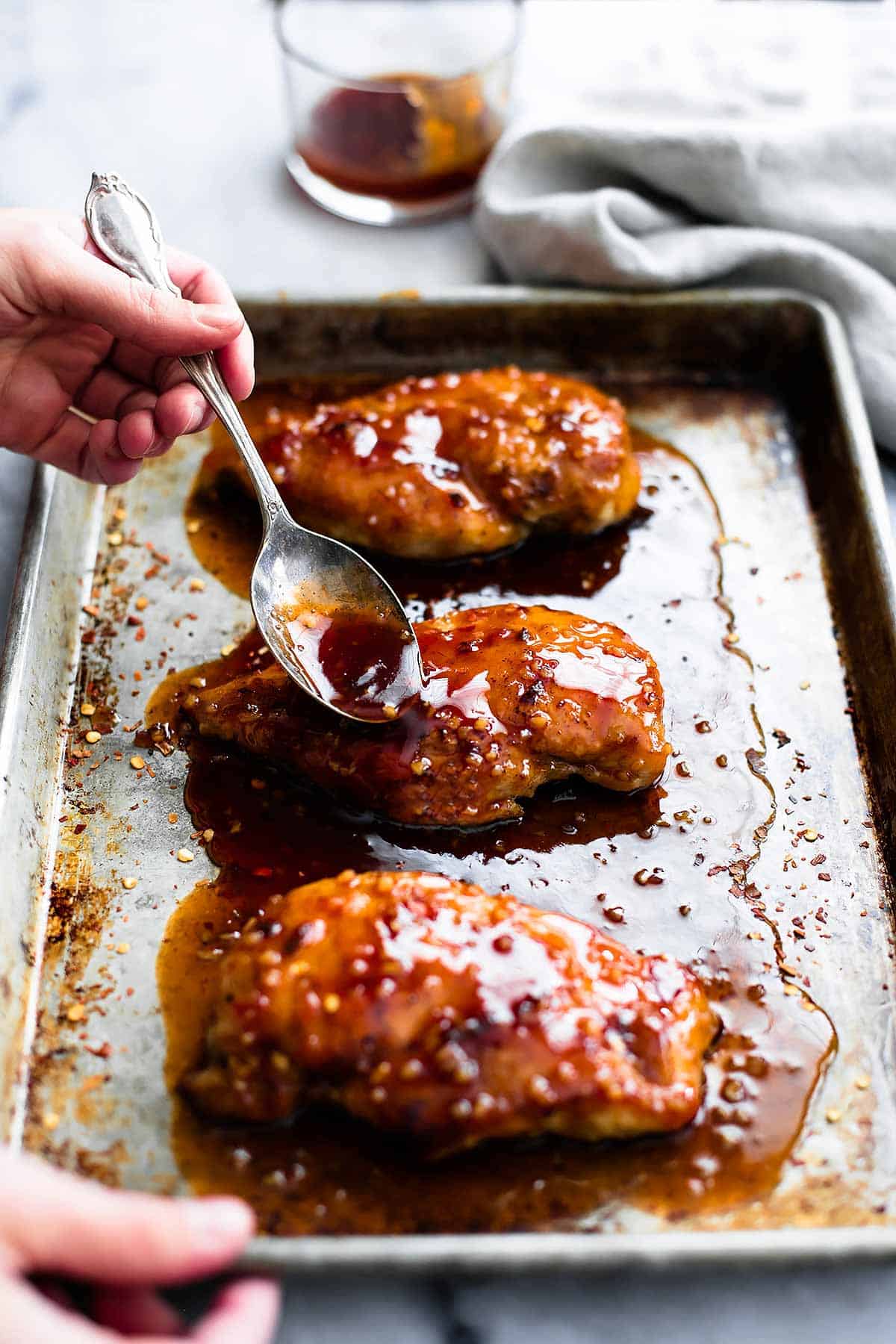 a hand pouring sauce on baked sweet & spicy chicken on a baking sheet with a spoon.