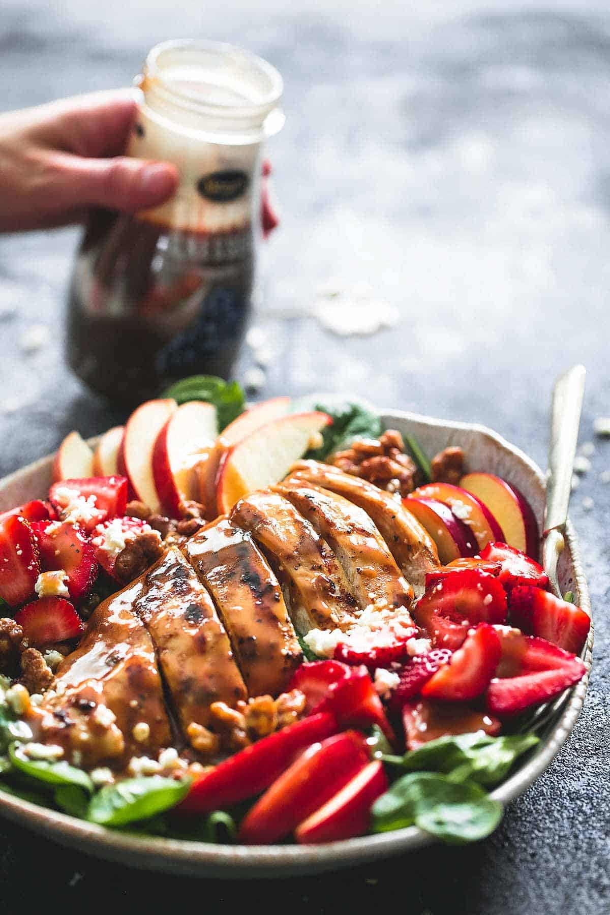balsamic chicken, strawberry, and apple salad and a fork on a plate with a hand holding a jar of balsamic dressing in the background.
