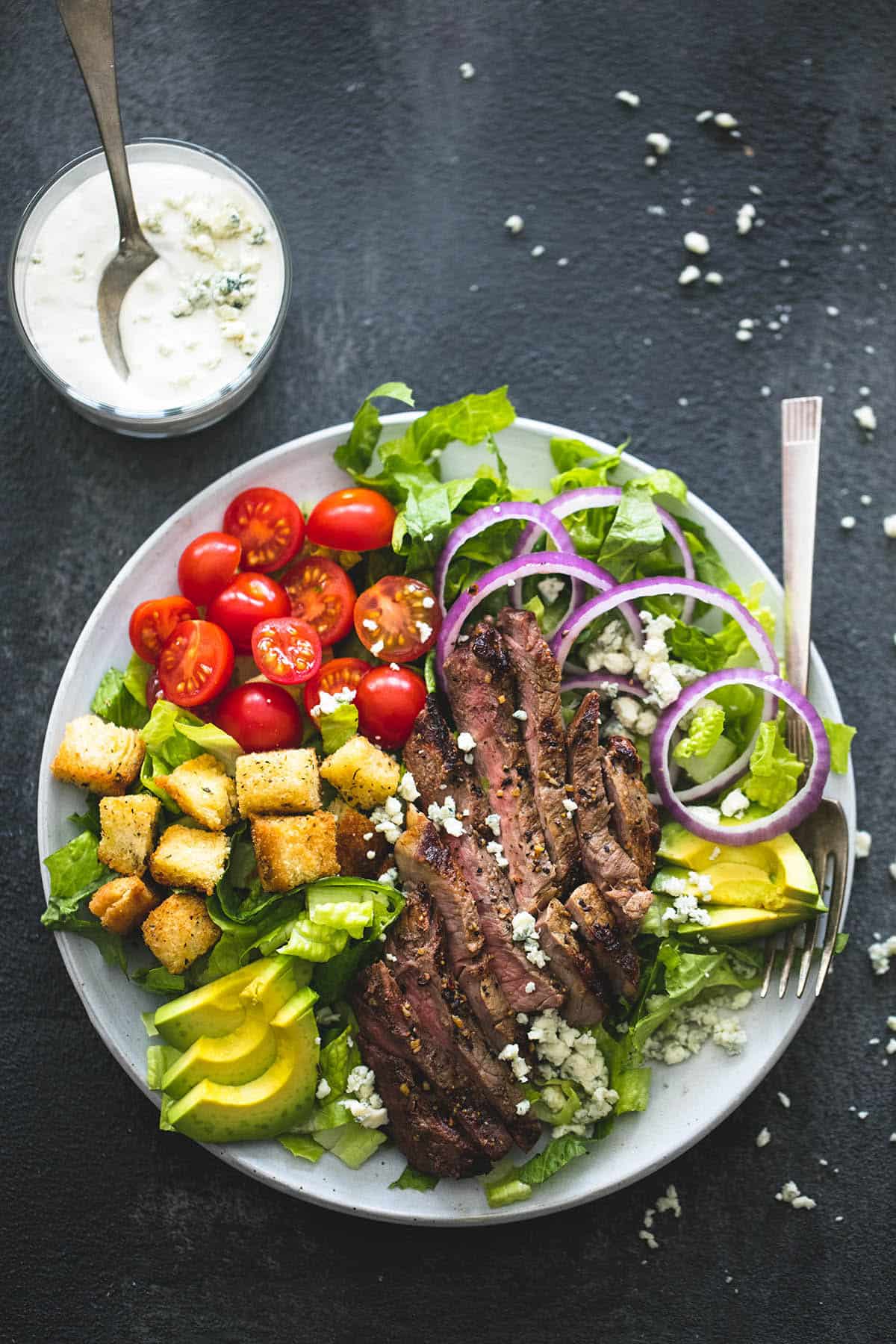 top view of black n' blue grilled steak salad with a fork on a plate with a glass of blue cheese dressing and a spoon on the side.