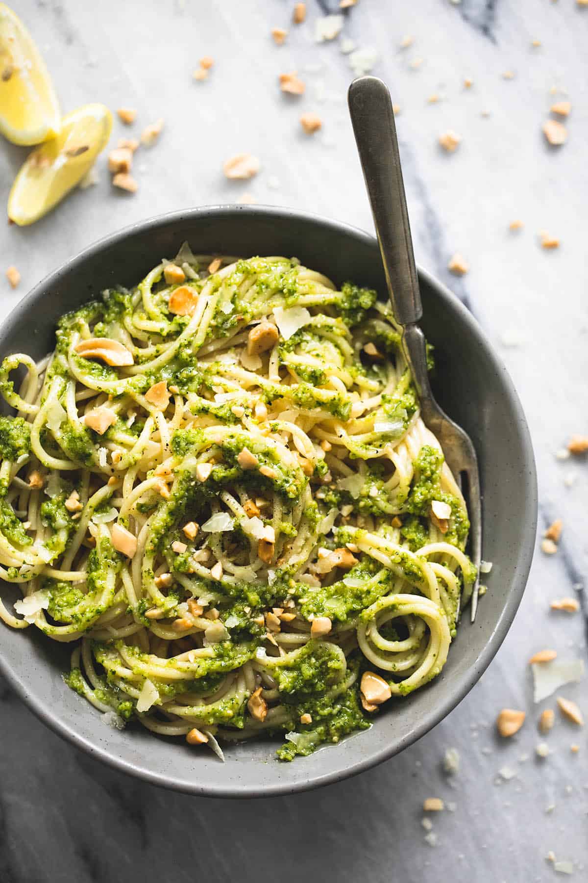 top view of cashew pesto pasta with a fork in a bowl with lemon slices on the side and crushed up cashews around the bowl.