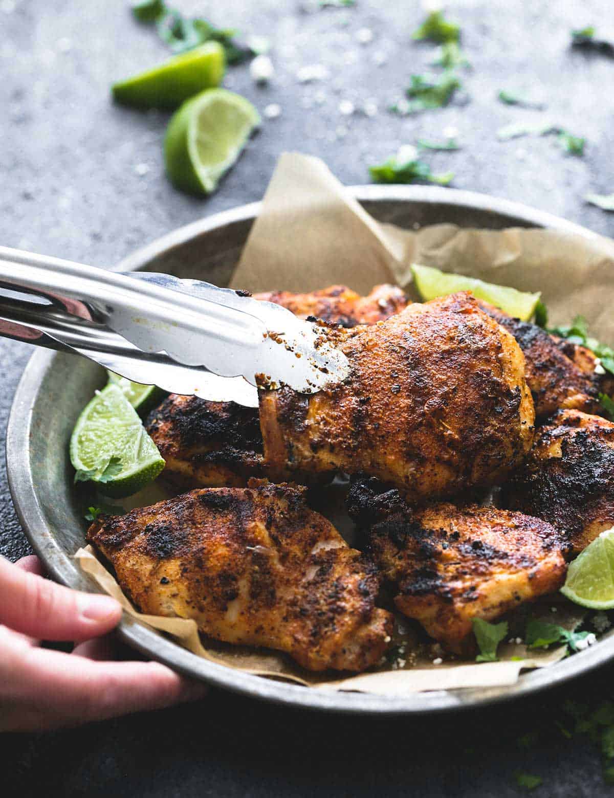 tongs picking up a piece of grilled Cajun lime chicken from a metal tray of chicken with lime slices in the background.