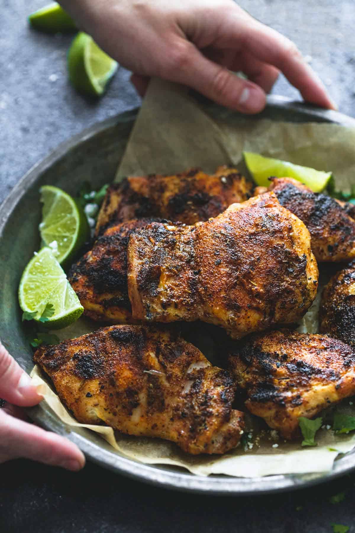 close up of hands holding a metal tray of grilled Cajun lime chicken with lime slices on the side.