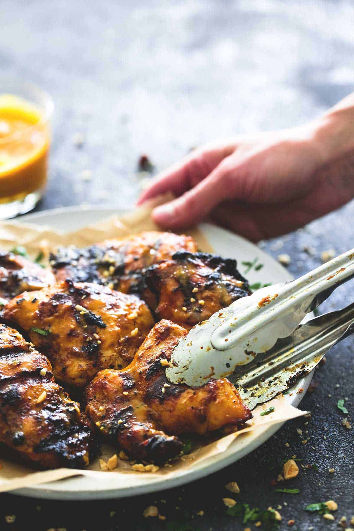 tongs picking up a piece of grilled Thai mango chicken from a plate of chicken with a hand holding one side of the plate.