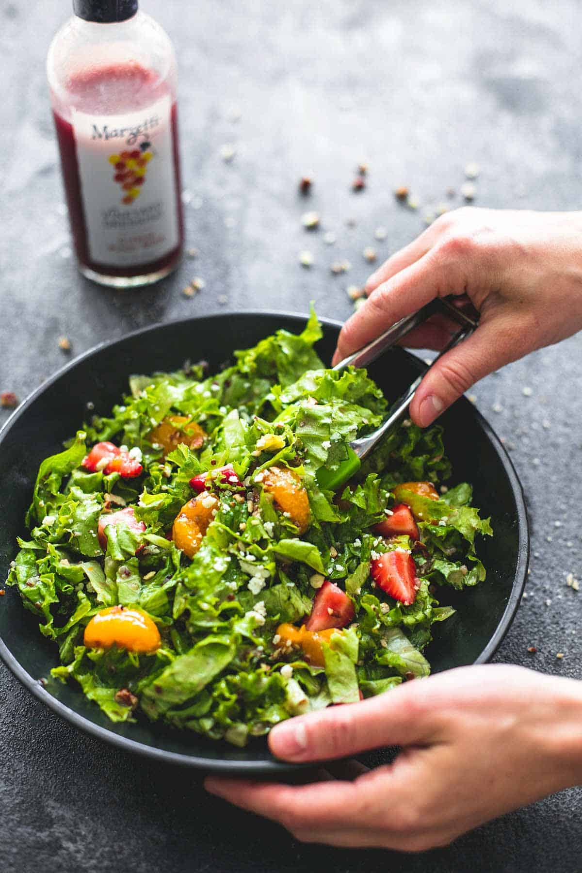 a pair of hands with one hand holding a bowl of mandarin strawberry quinoa chopped salad and the other tossing the salad with a container of berry vinaigrette on the side.
