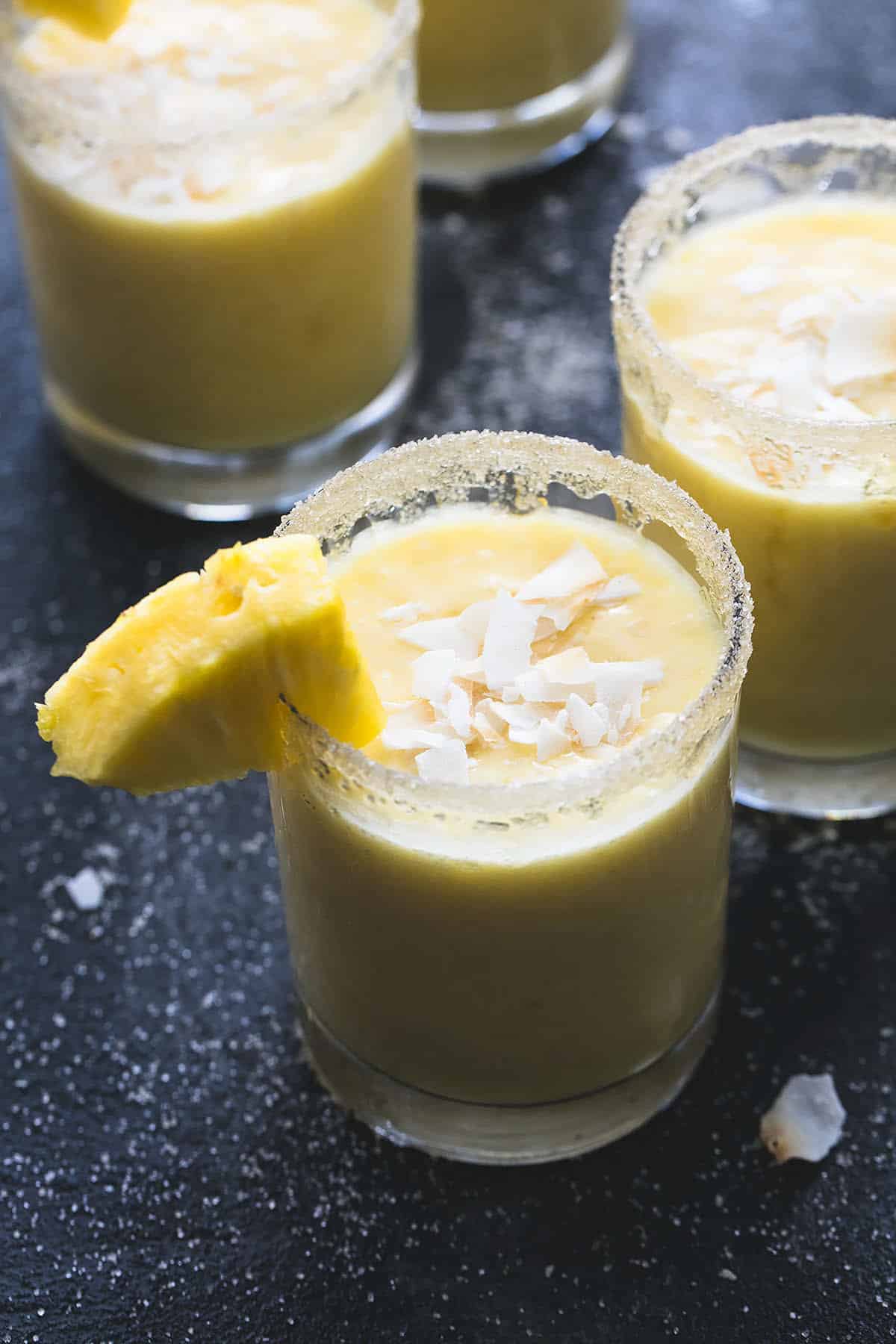 a glass of a pina colada smoothie with coconut shavings on top and a chunk of pineapple on the side with more glasses of smoothie in the background.