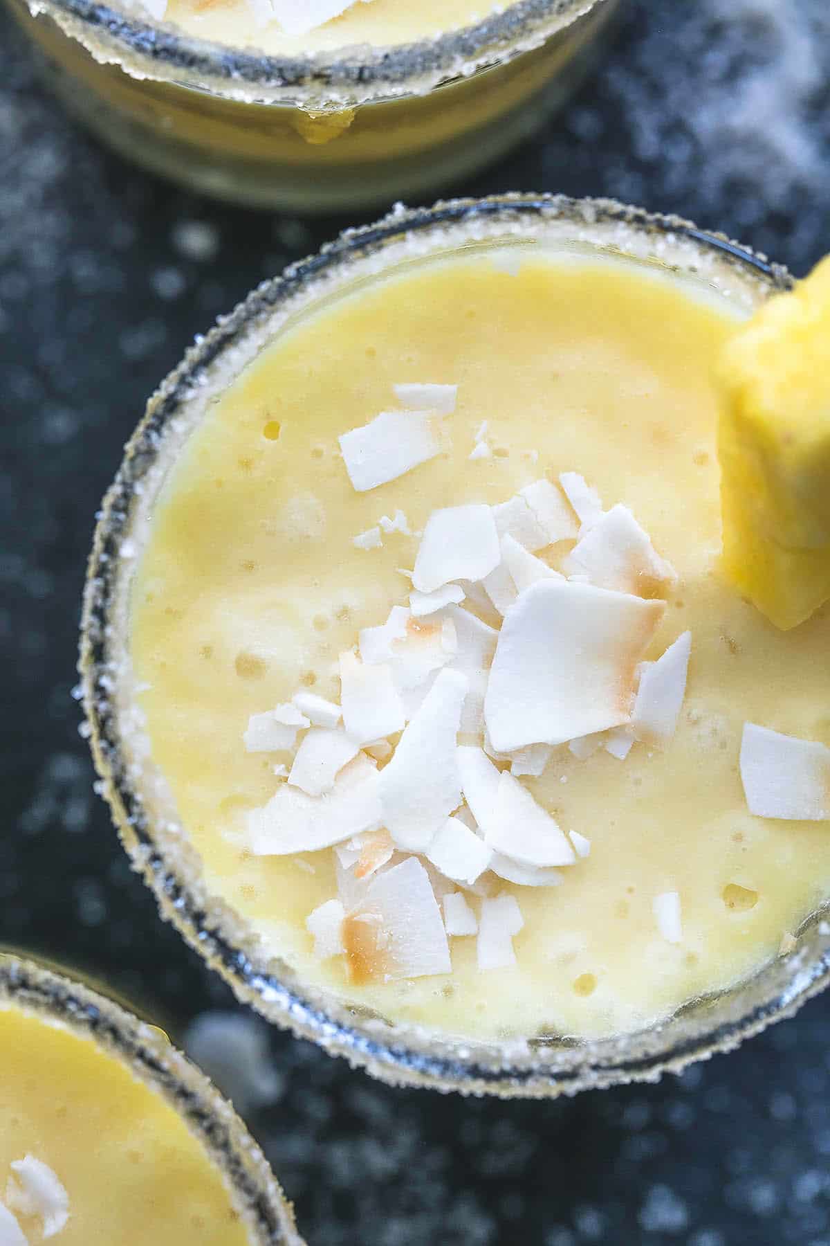 close up top view of a pina colada smoothie in a glass with coconut shavings on top and a pineapple chunk on the side with more smoothies in glasses on the side.