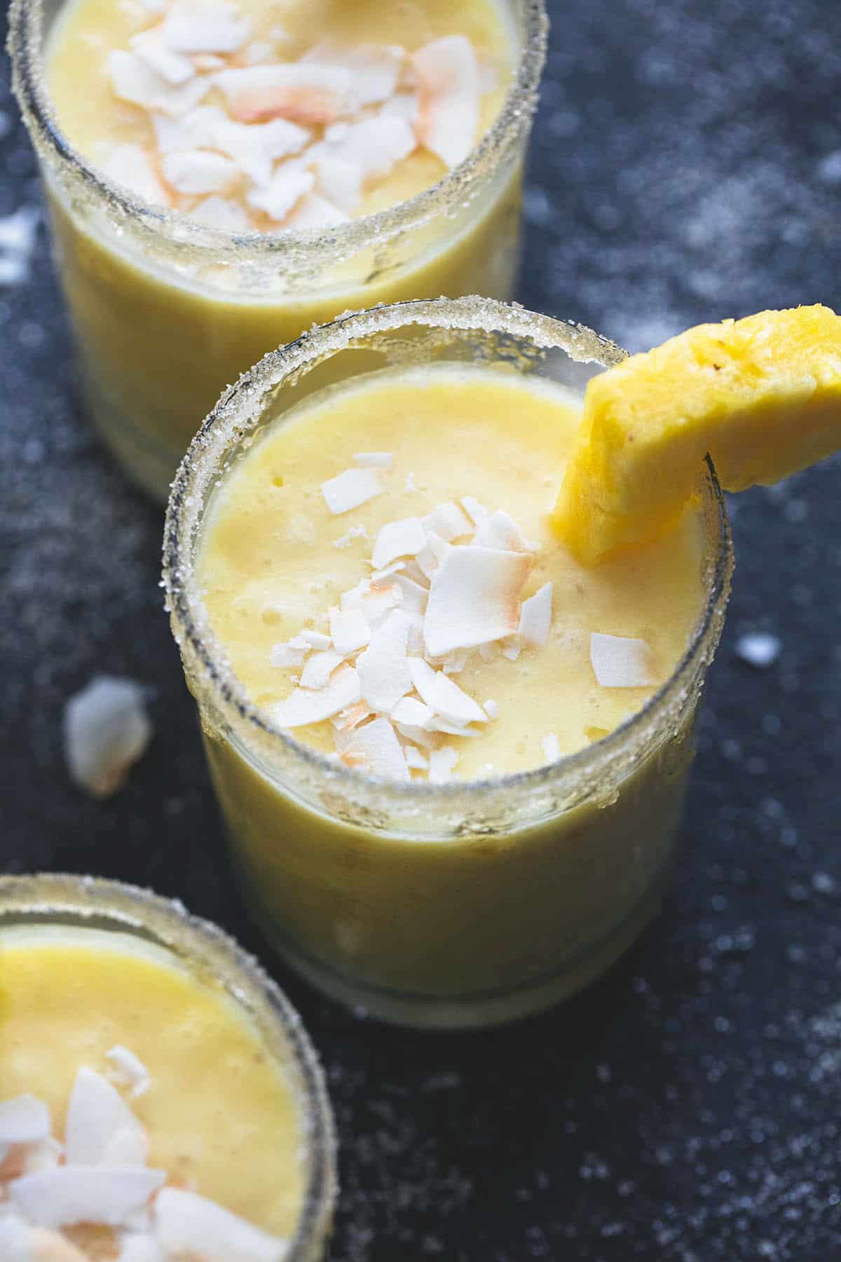 a pina colada smoothie in a glass with coconut shavings on top with a pineapple chunk on the side with more smoothies in glasses on the side.