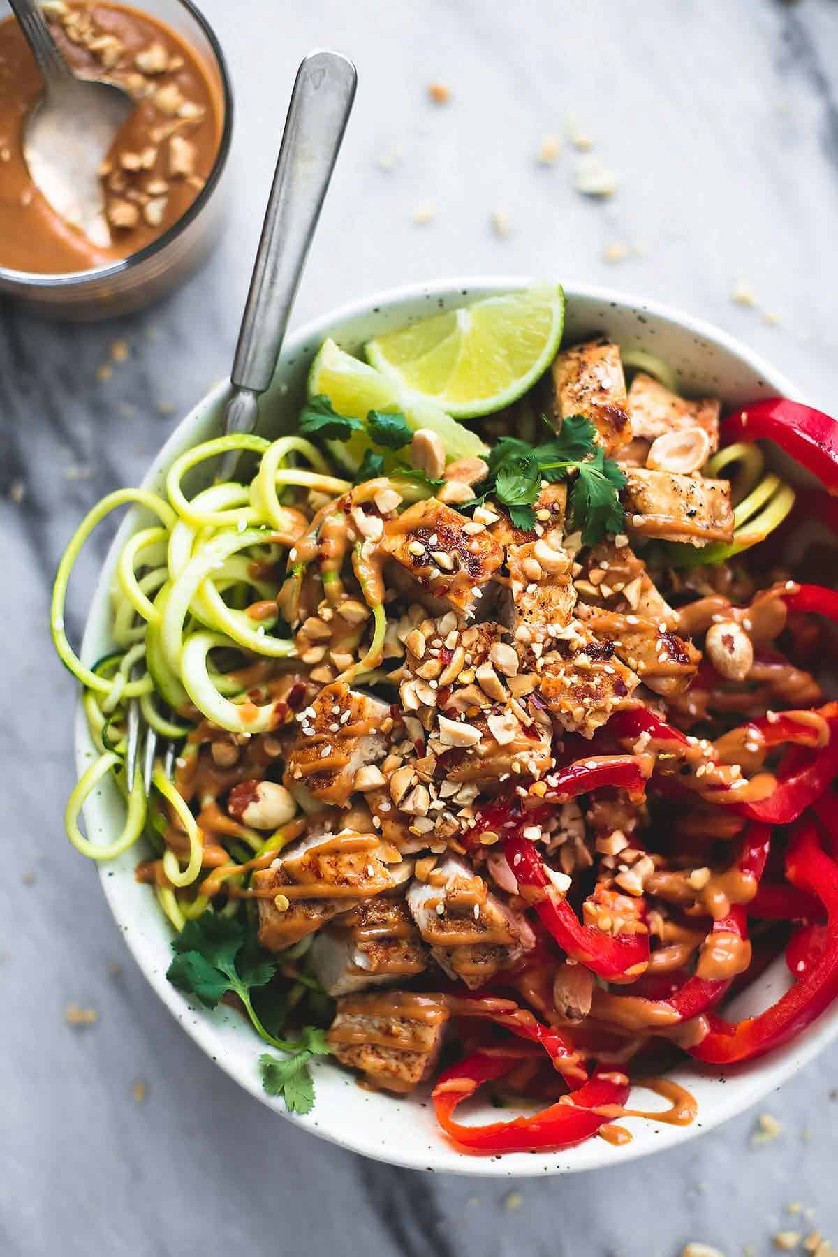 top view of a Thai peanut chicken & zucchini noodle bowl with a fork and a glass of peanut sauce with a spoon on the side.