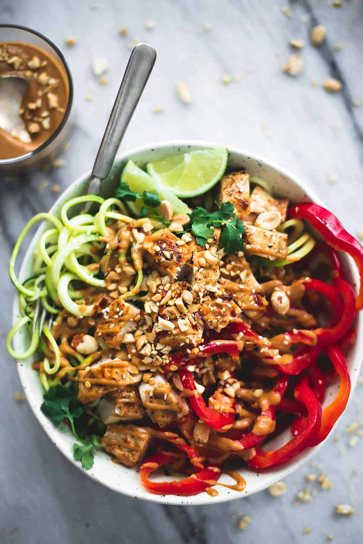 top view of a Thai peanut chicken & zucchini noodle bowl with a fork and a glass of peanut sauce on the side.