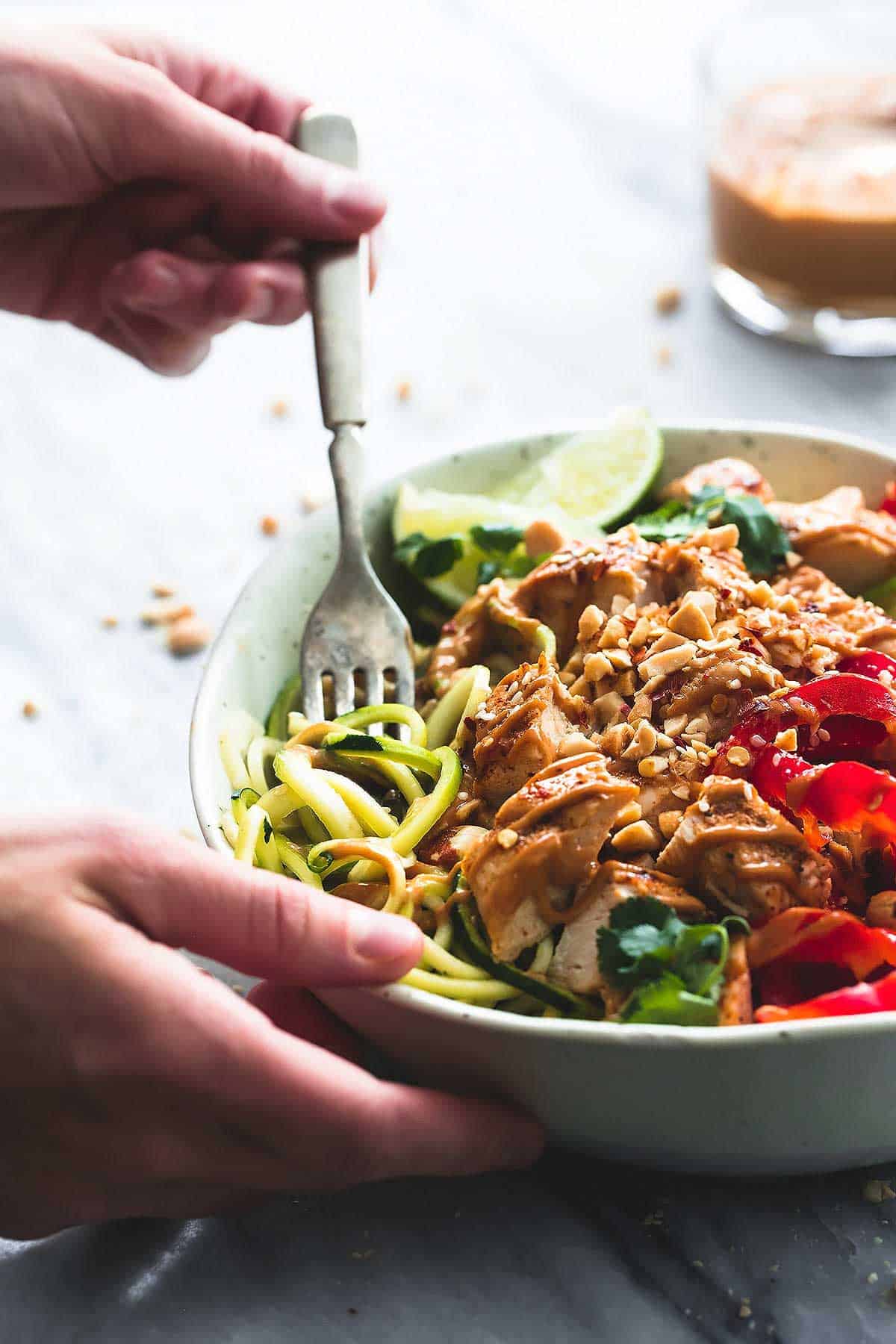 a hand holding a fork in a Thai peanut chicken & zucchini noodle bowl with a glass of peanut sauce in the background.