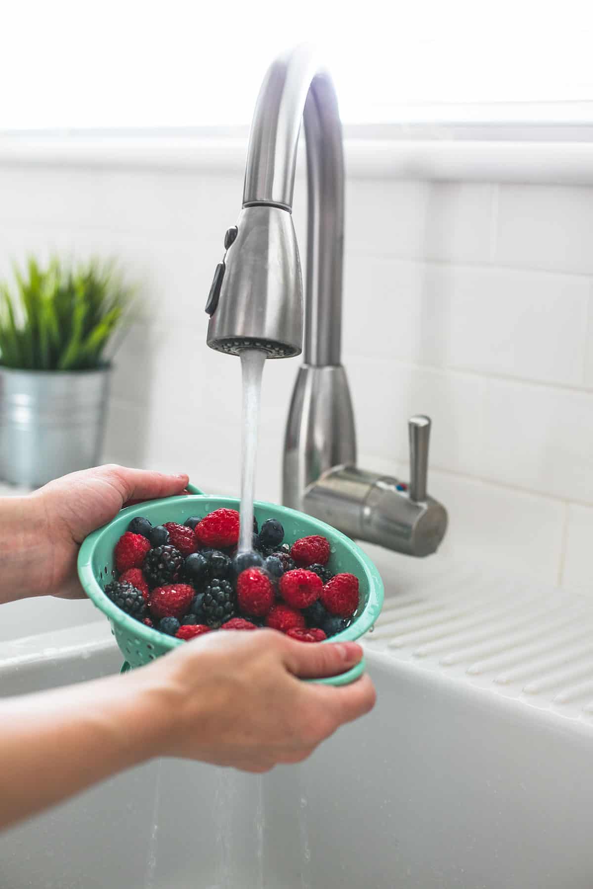 a hand holding a small colander full of mixed berries under a running faucet.