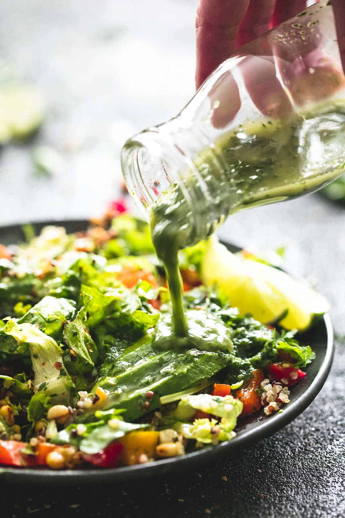 a hand pouring a jar of cilantro lime dressing on top of chopped fiesta quinoa salad on a plate.