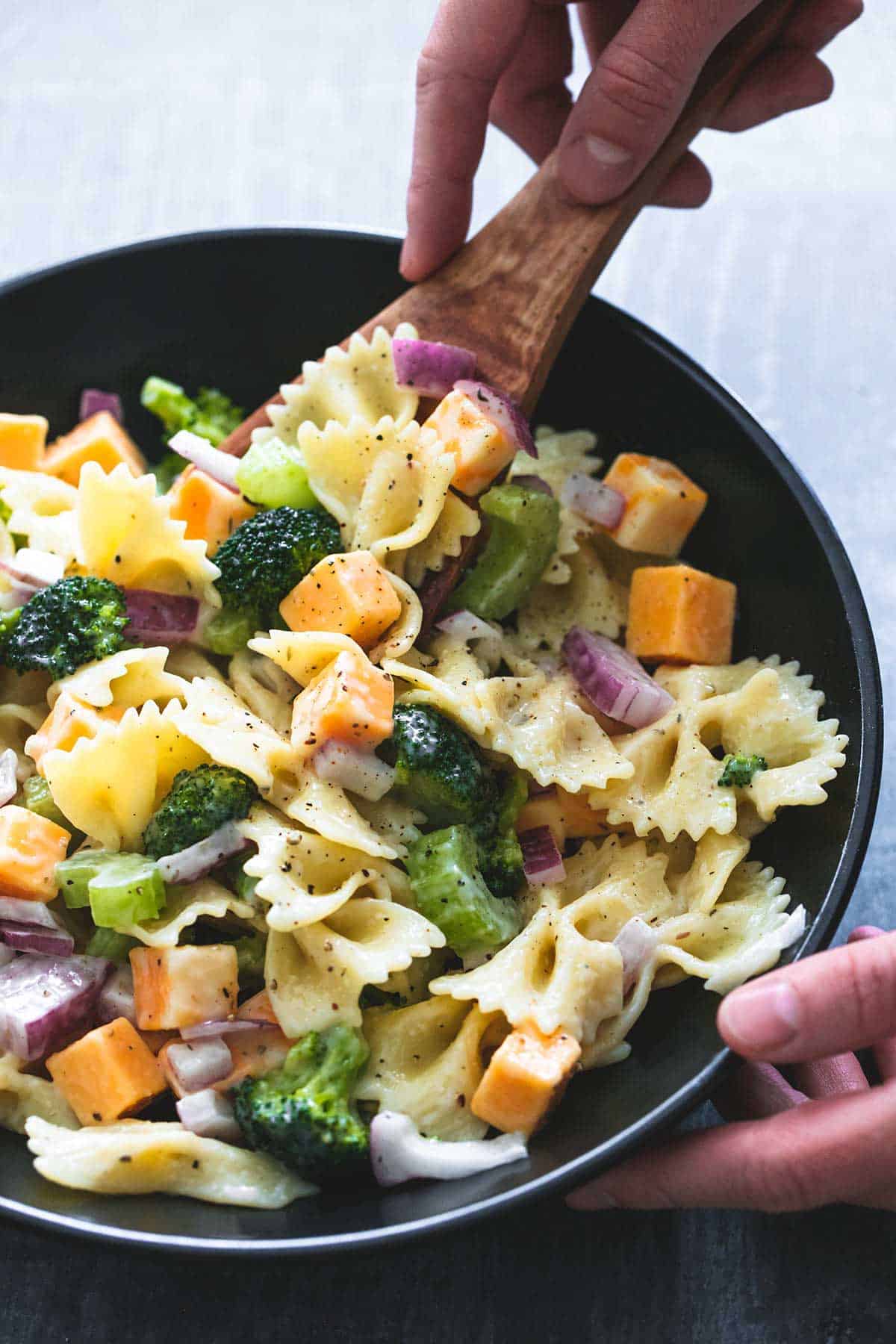 a hand scooping some creamy cheddar broccoli salad from a bowl with a wooden serving spoon.
