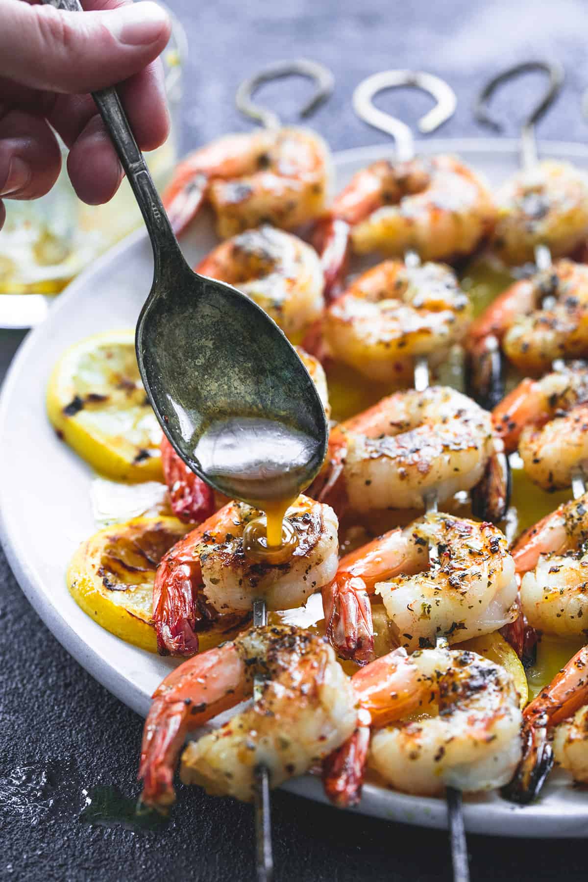 a hand pouring some lemon butter mixture on grilled lemon garlic shrimp on skewers on a plate with a spoon.