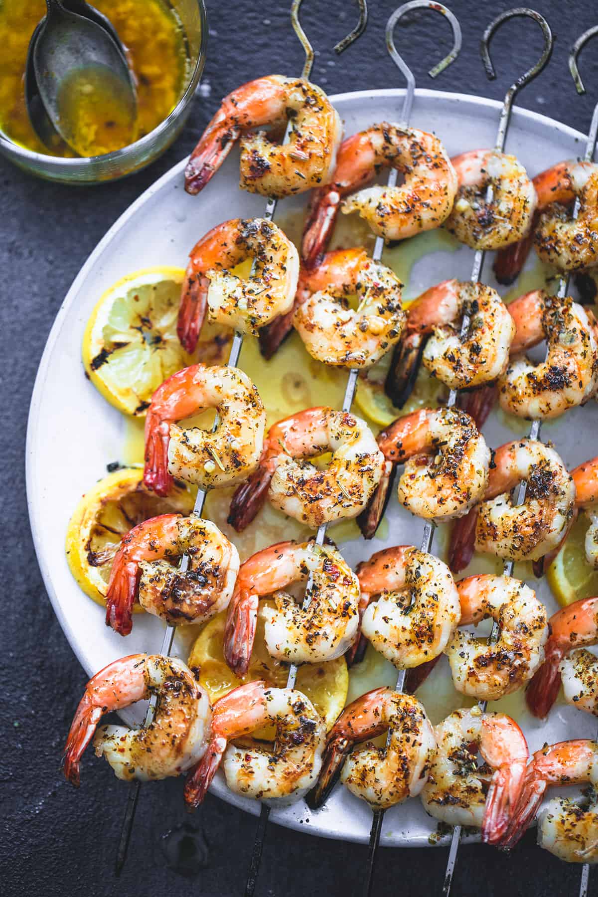 top view of grilled lemon garlic shrimp on skewers on a plate with lemon butter mixture and a spoon in a glass on the side.
