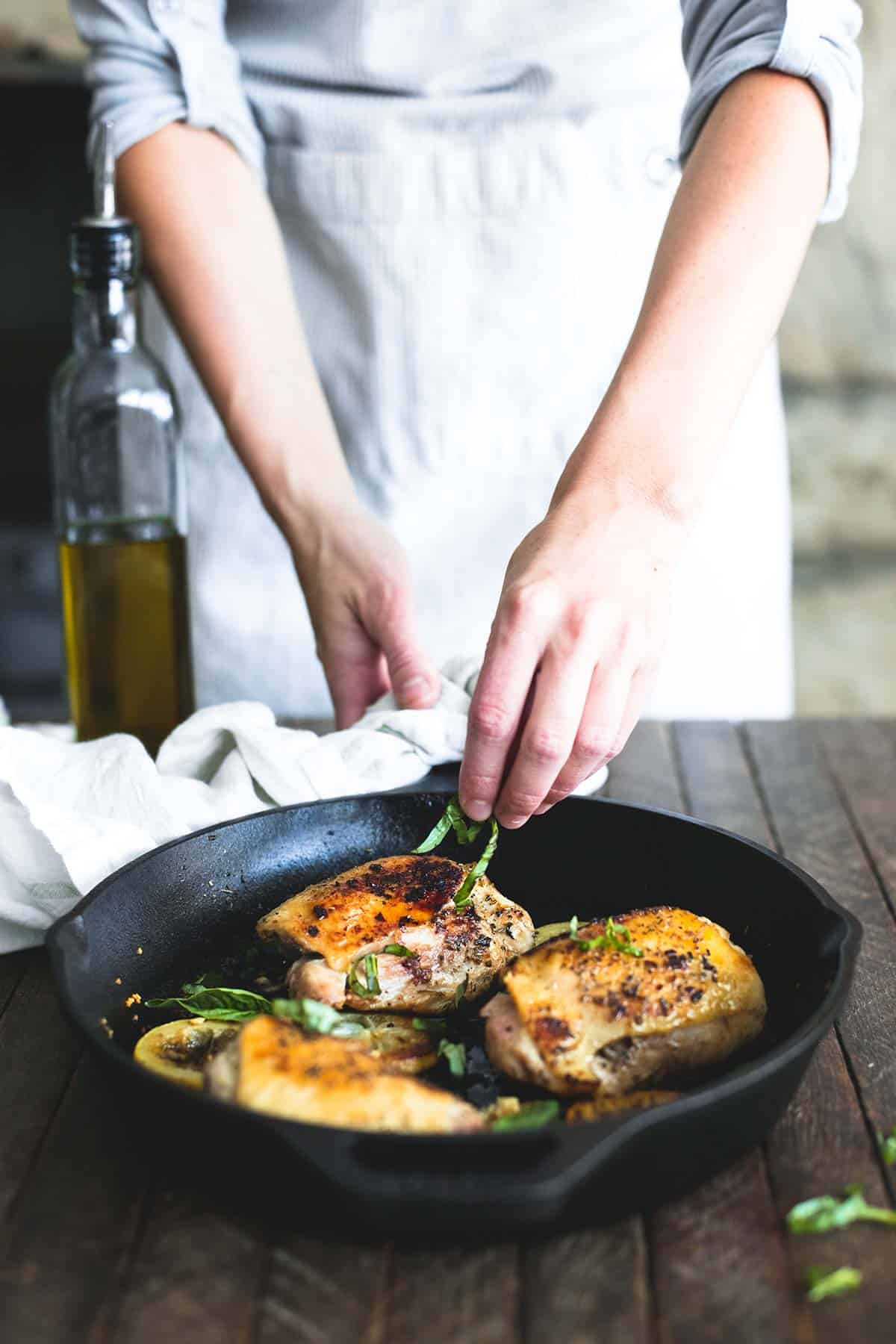 someone putting garnish on top of buttery lemon herb chicken thighs in a skillet with a bottle of oil on the side.