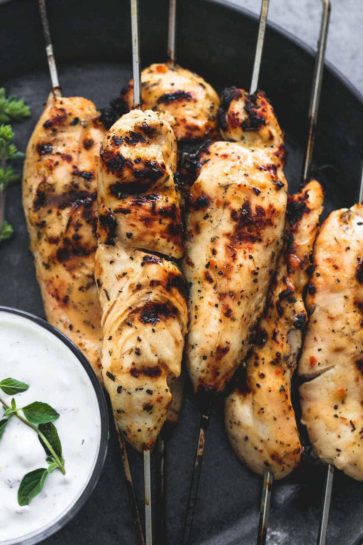 close up top view of grilled chicken souvlaki on skewers with tzatziki sauce in a bowl on the side all on a plate.