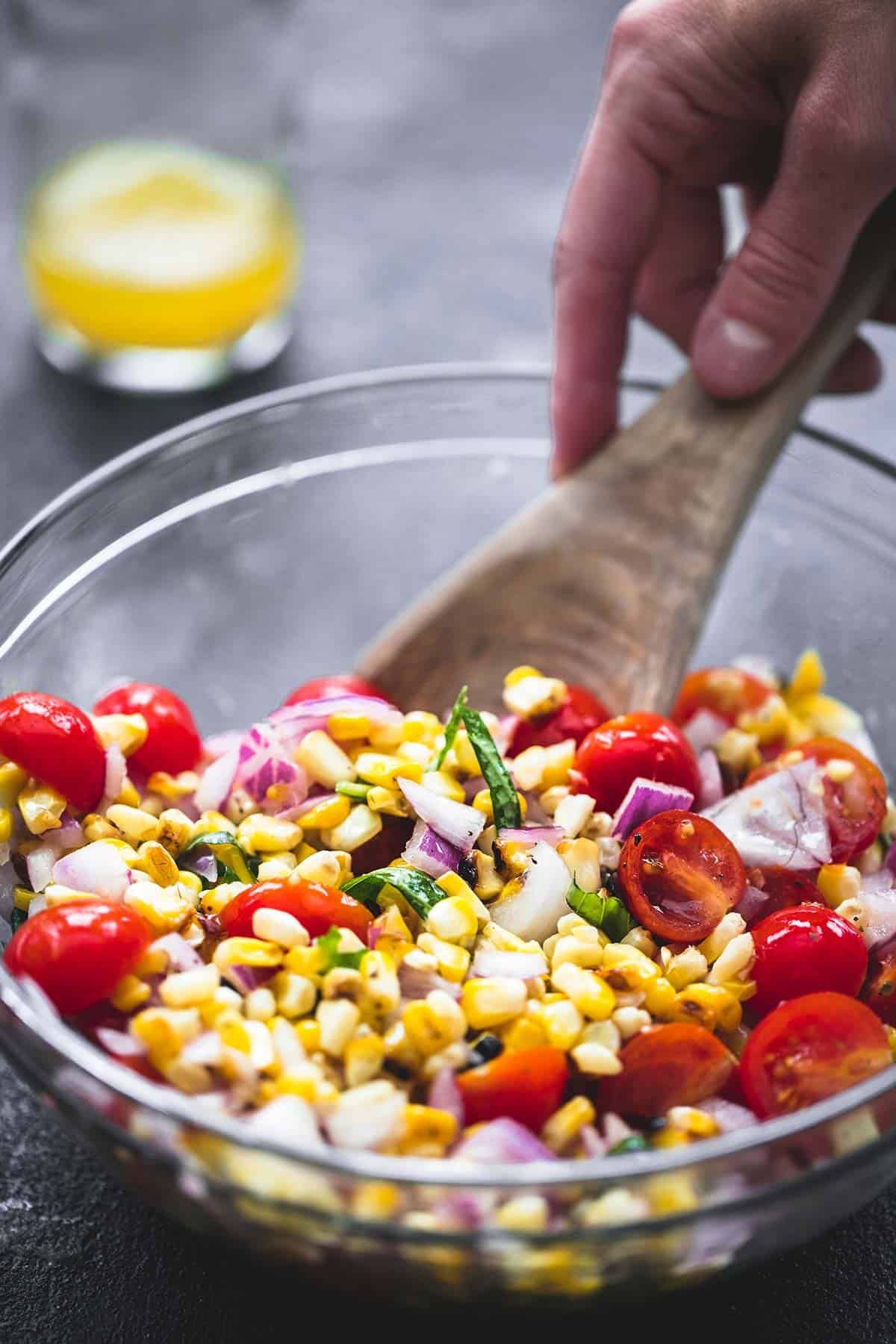 a hand scooping up some grilled corn salad from a bowl with a wooden serving spoon.