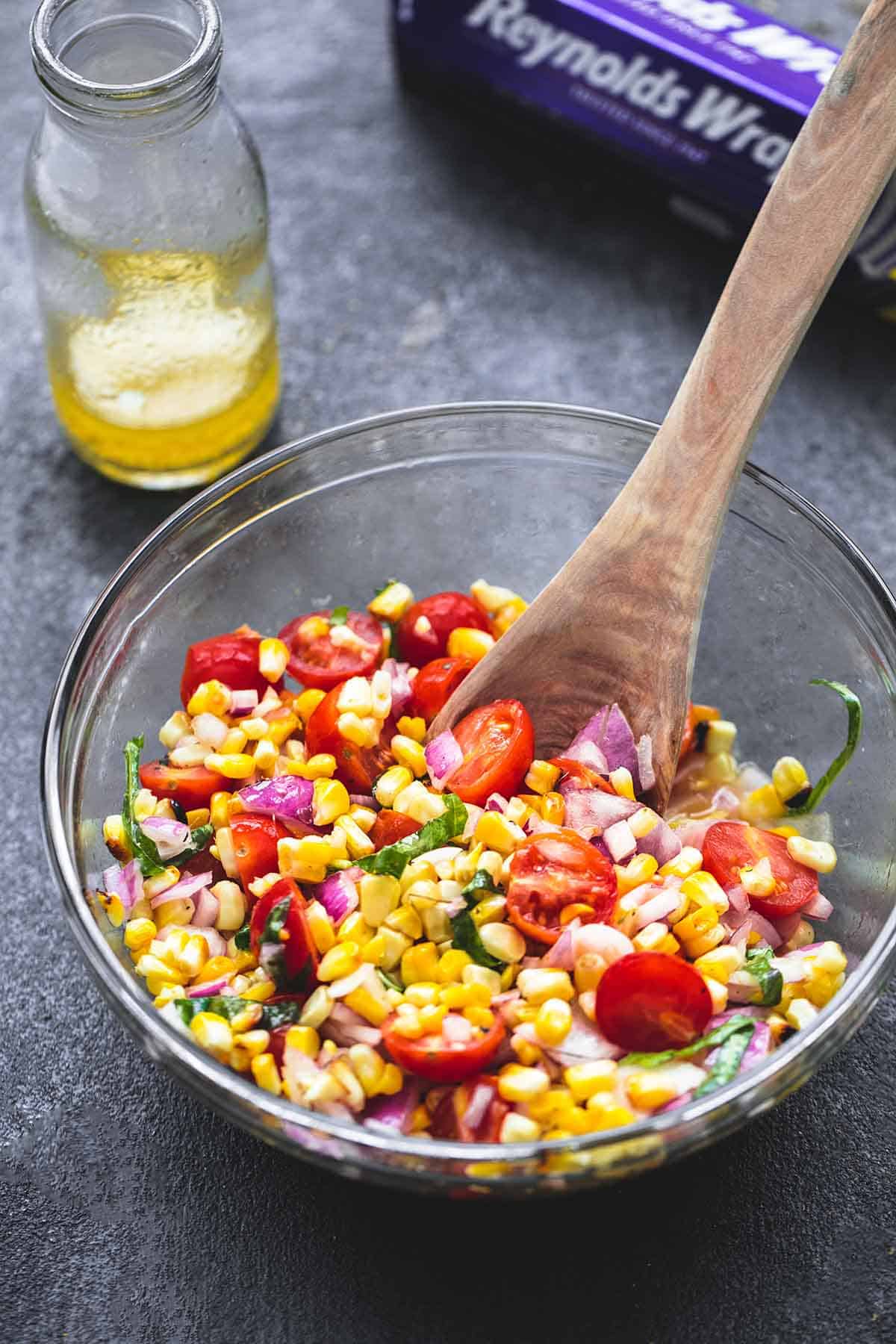 grilled corn salad with a wooden serving spoon in a bowl with a jar of dressing and an aluminum foil box on the side.