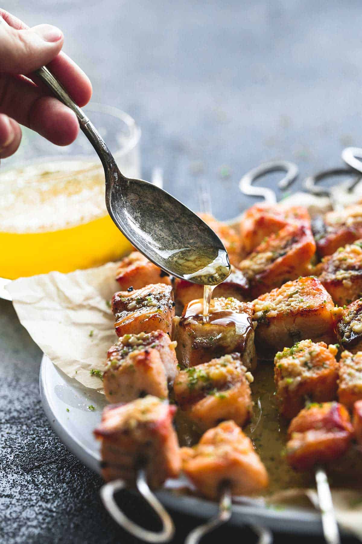 a hand pouring some butter sauce on top of grilled lime butter salmon skewers on a plate with a spoon with more sauce in a glass on the side.