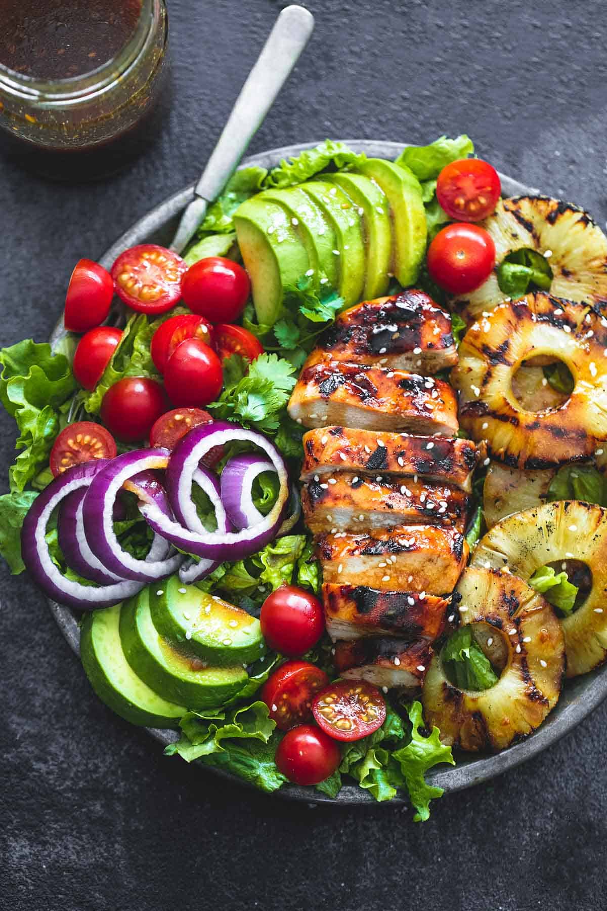 grilled teriyaki chicken salad with a fork on a plate with teriyaki vinaigrette in a jar on the side.