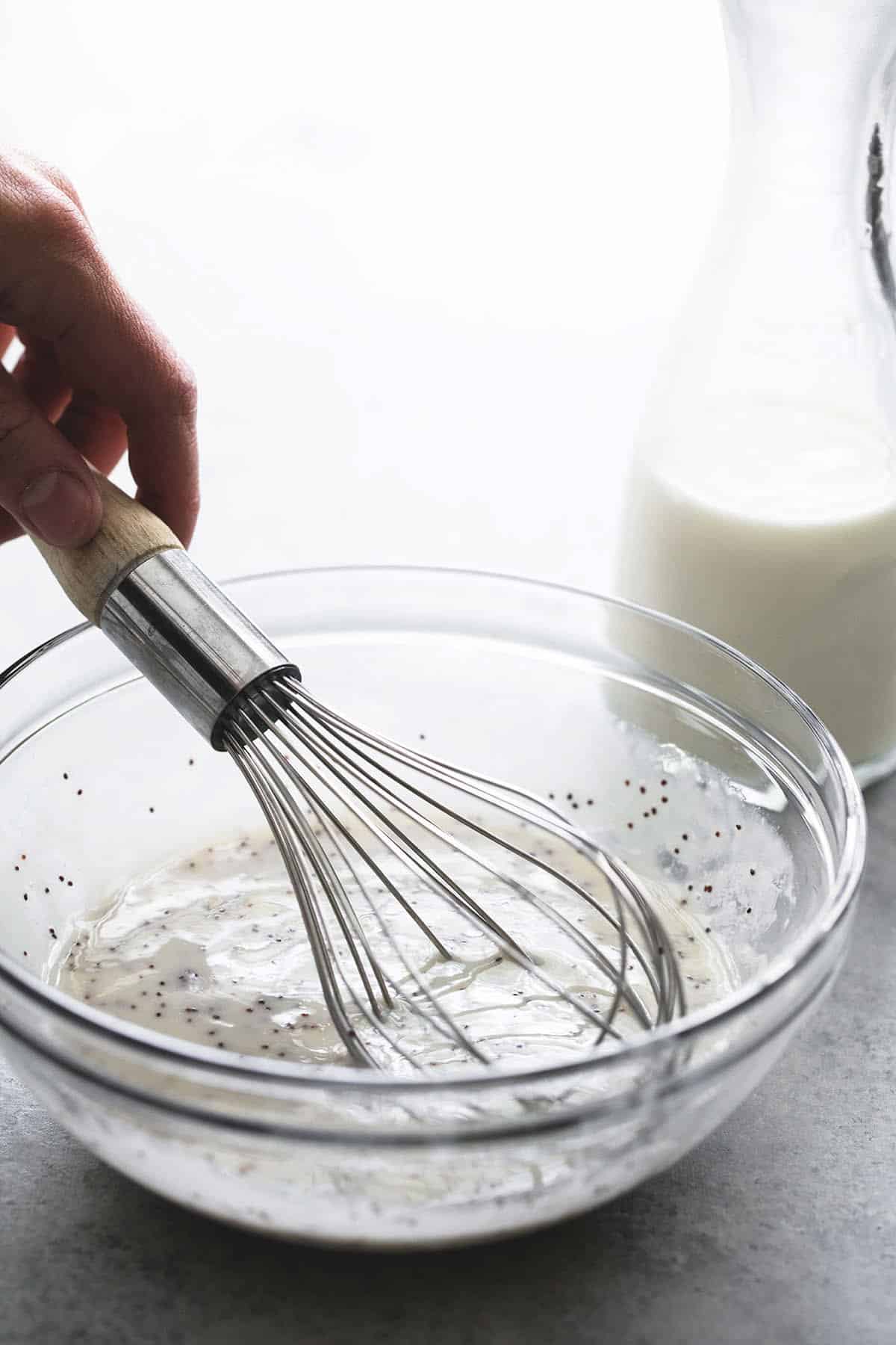 a hand mixing honey lime dressing with a whisk in a bowl with a milk in a carafe on the side.