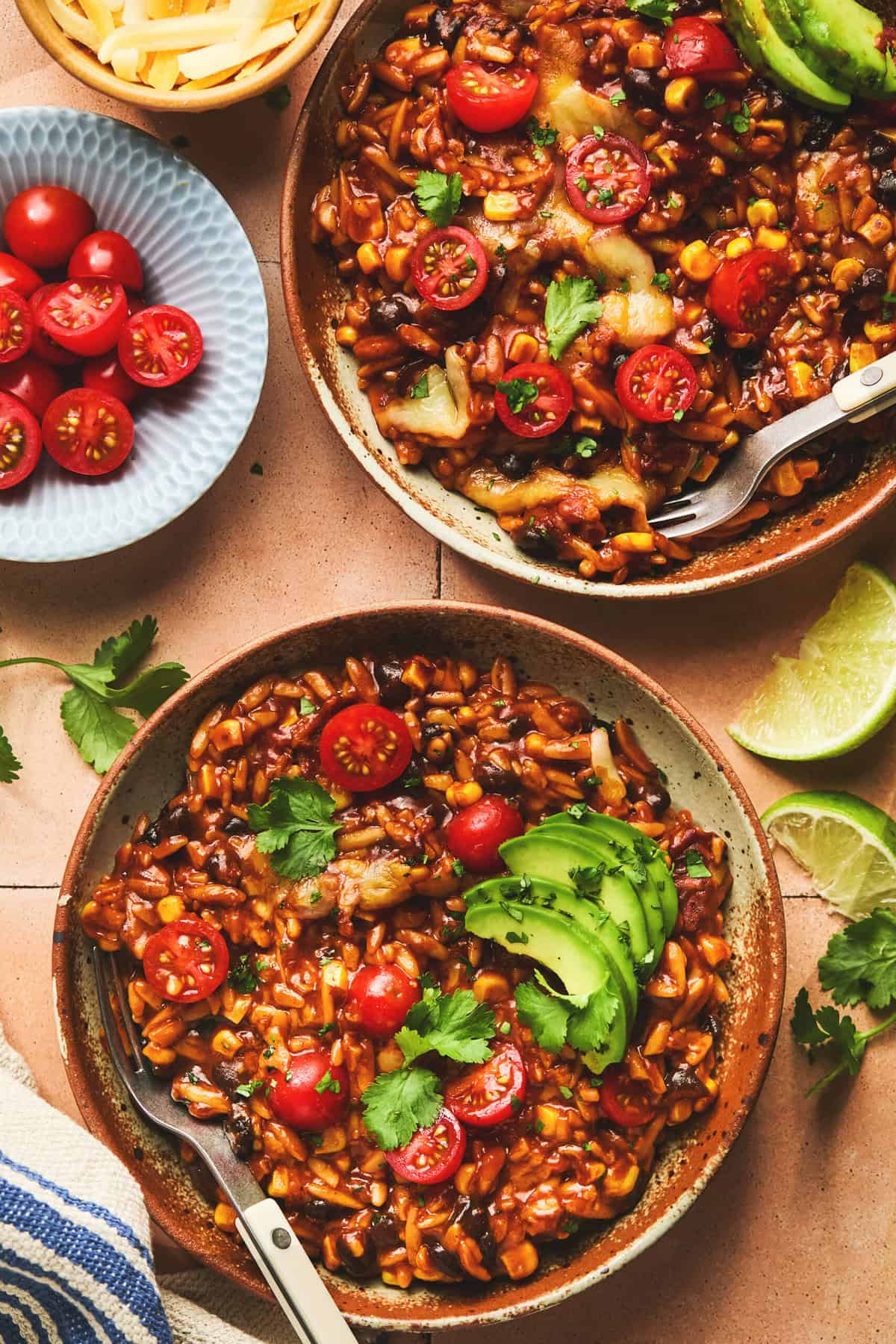 two bowls of cooked orzo in enchilada sauce with cheese, tomatoes, and avocado slices.