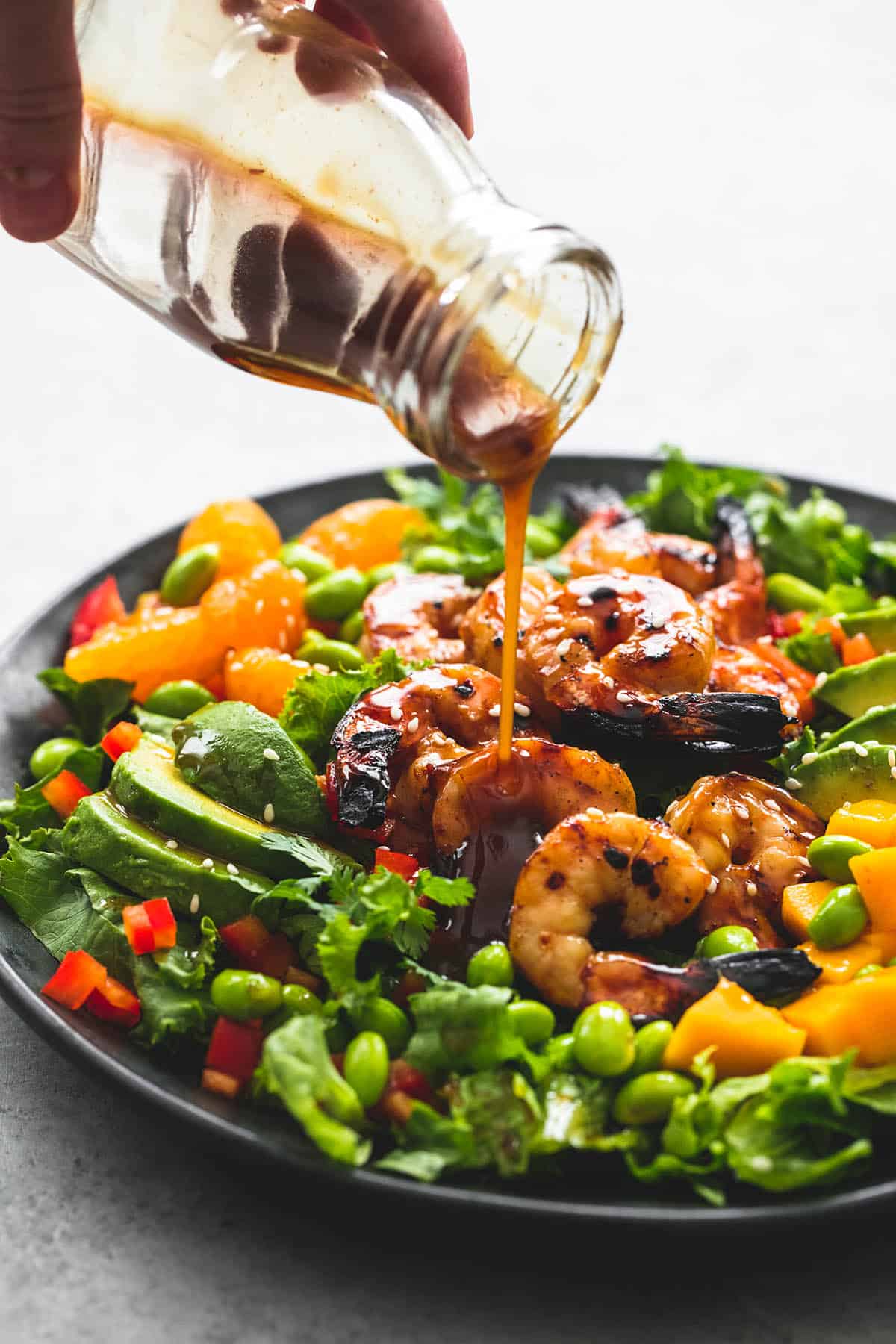 a hand pouring dressing on mango mandarin sesame shrimp salad on a plate.