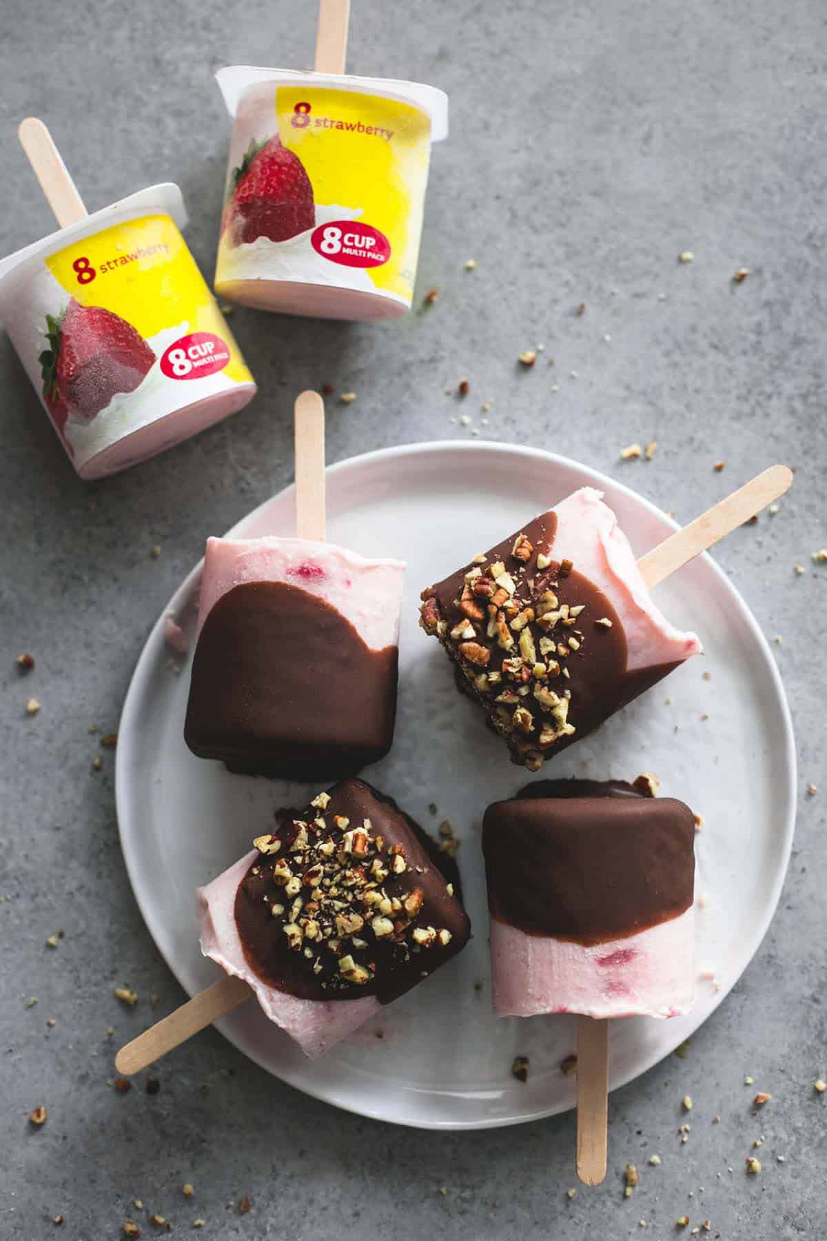 top view of chocolate covered strawberry yogurt pops on a plate with some topped with crushed pecans with two yogurt containers laying on their sides with popsicle sticks in them on the side.