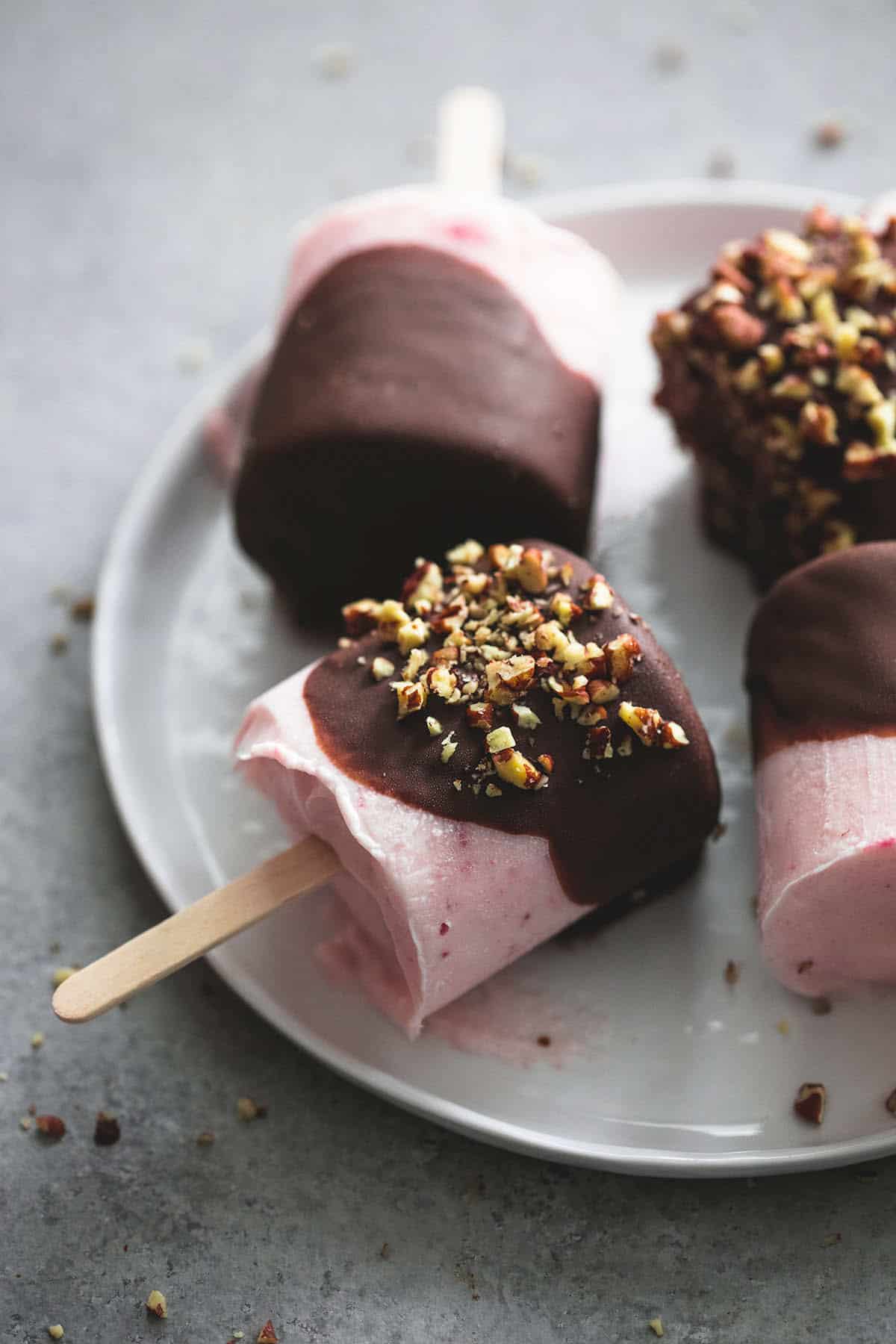 close up of chocolate covered strawberry yogurt pops on a plate with a couple topped with crushed pecans.