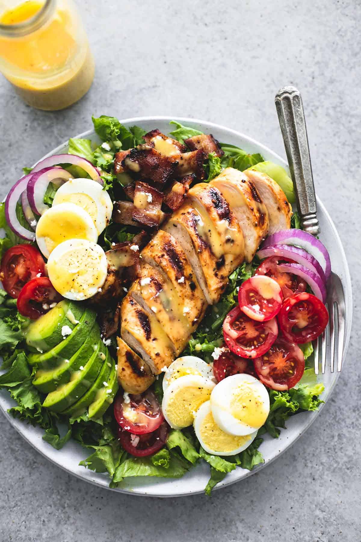 top view of honey mustard chicken cobb salad and a fork on a plate with a jar of dressing on the side.