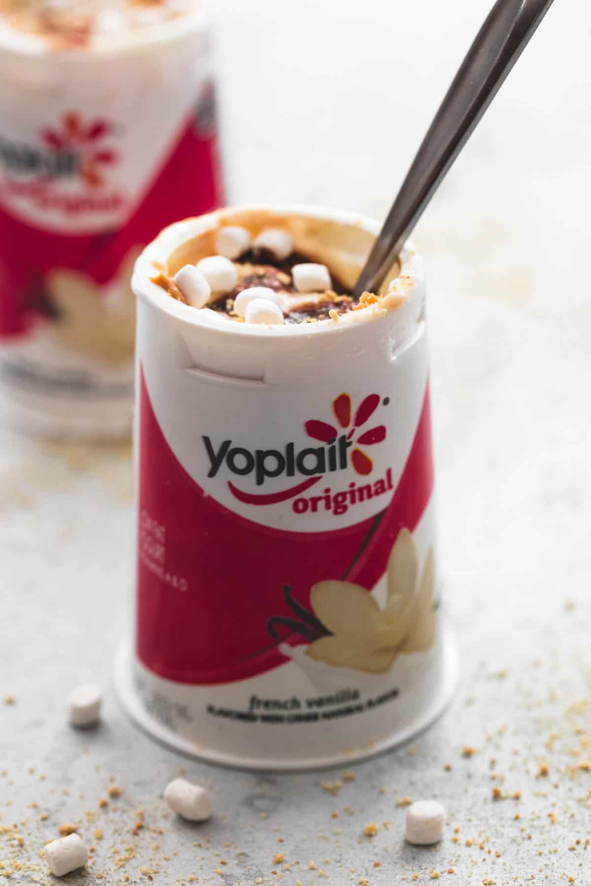 peanut butter s'mores yogurt with a spoon in a Yoplait container with another yogurt container faded in the background.