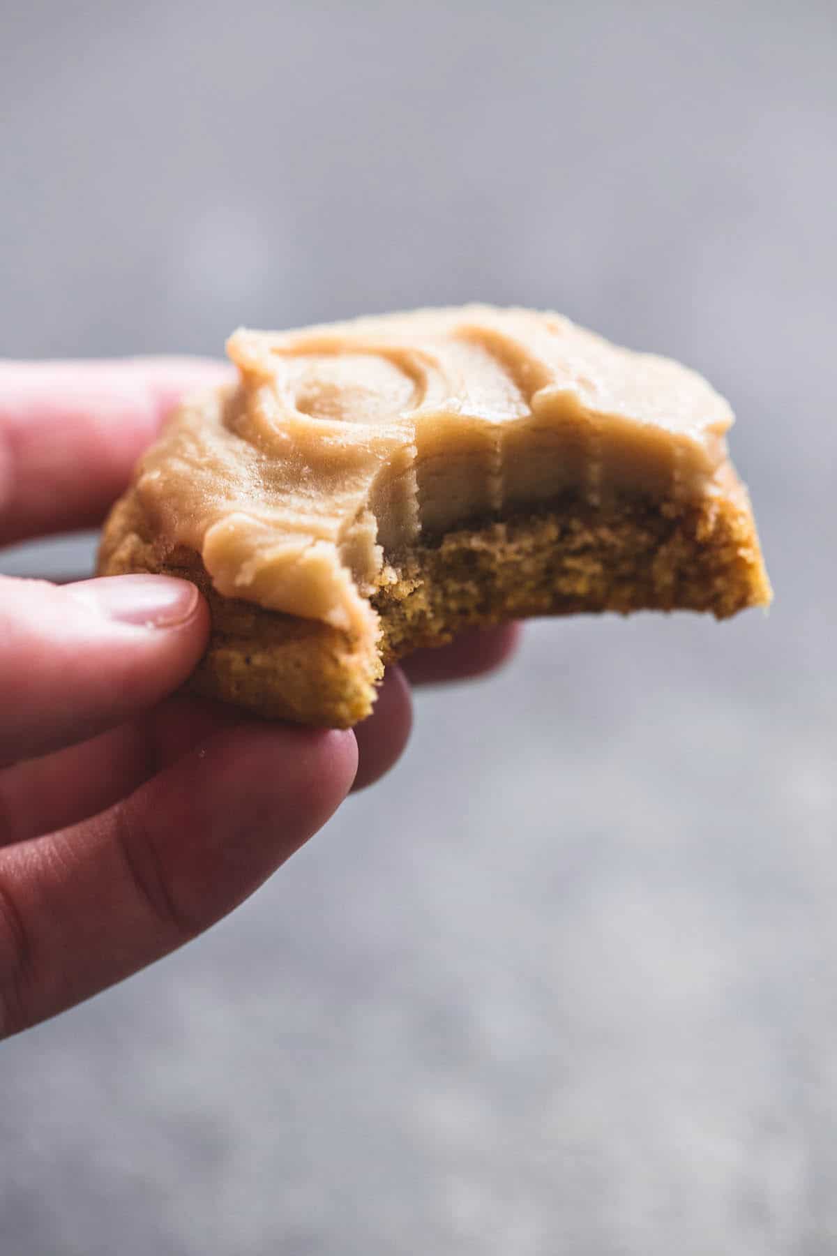 a hand holding a pumpkin cookie with brown sugar frosting with a bite missing.