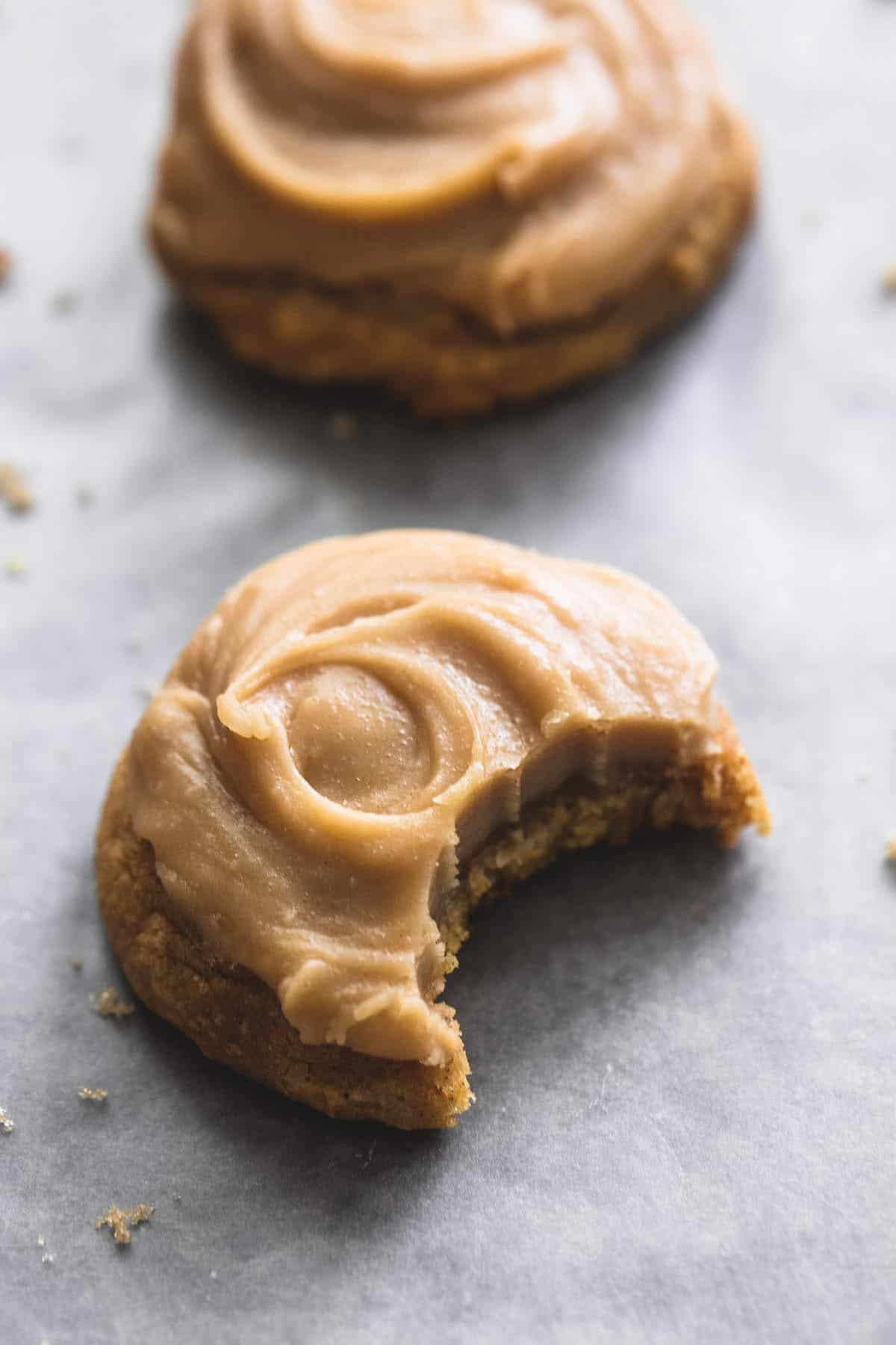 a pumpkin cookie with brown sugar frosting with a bite missing with another cookie in the background.