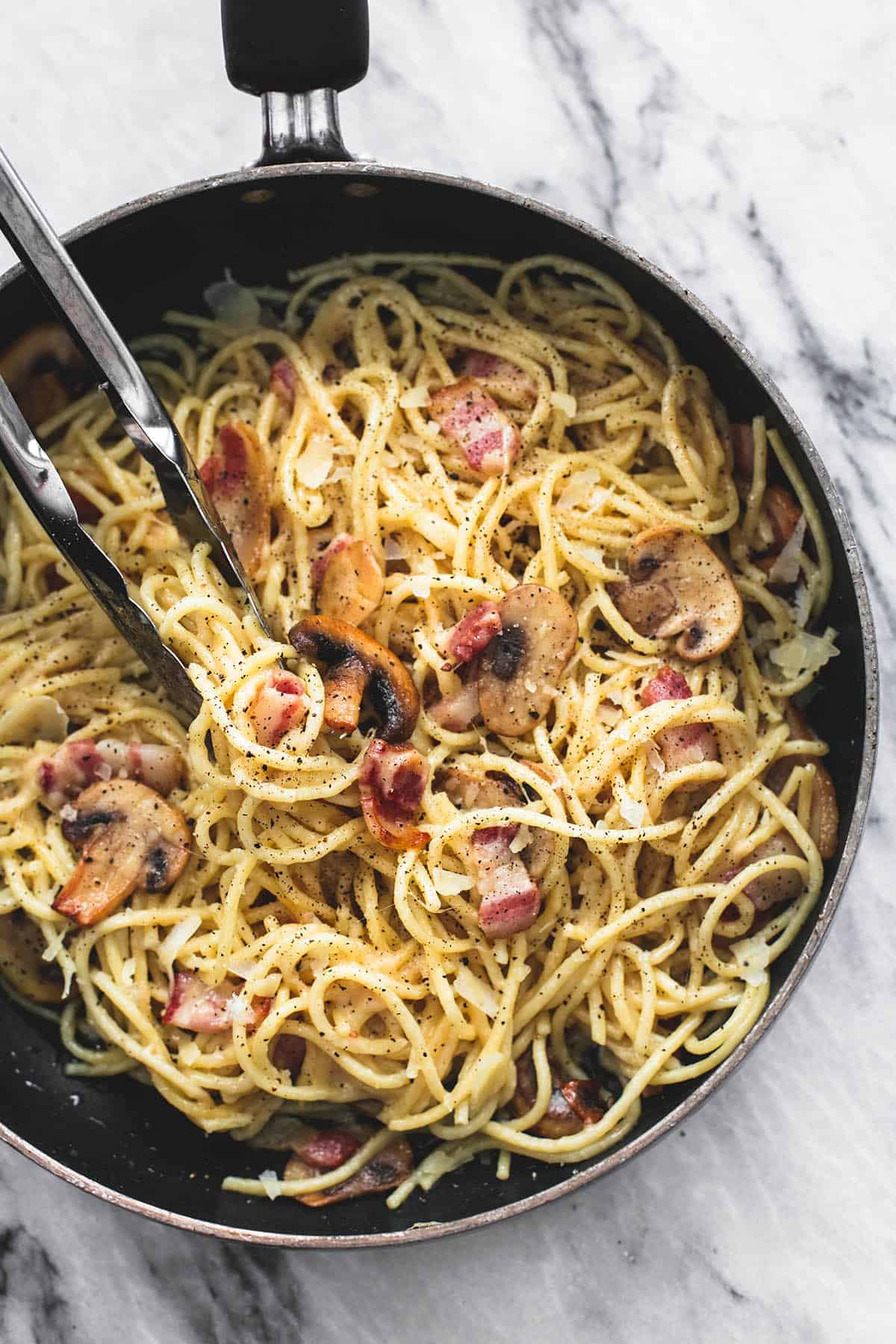 top view of cacio-e-pepe with mushrooms and bacon with tongs in a pan.