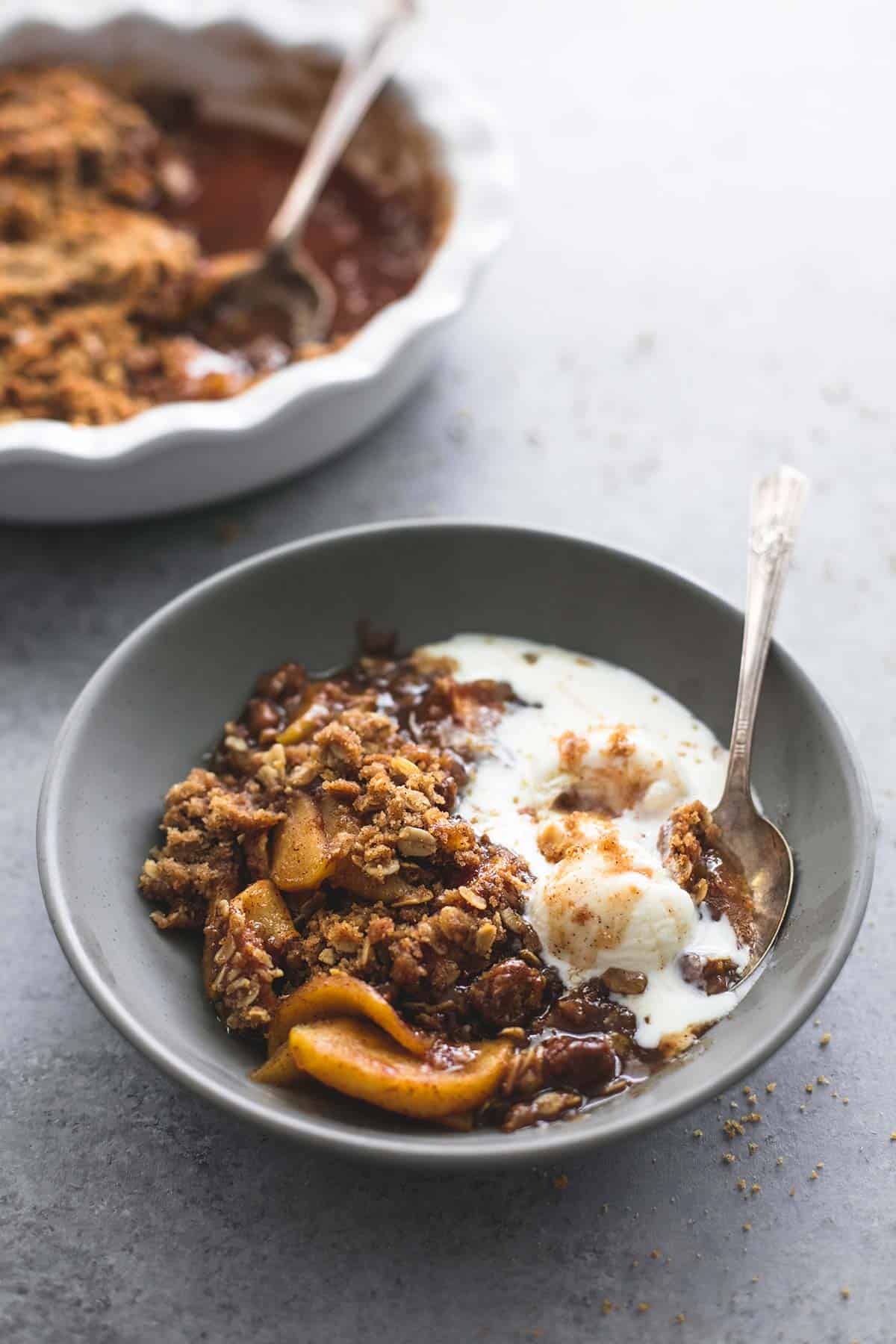 apple crisp with vanilla ice cream and a spoon in a bowl with more apple crisp and a serving spoon in a serving bowl.