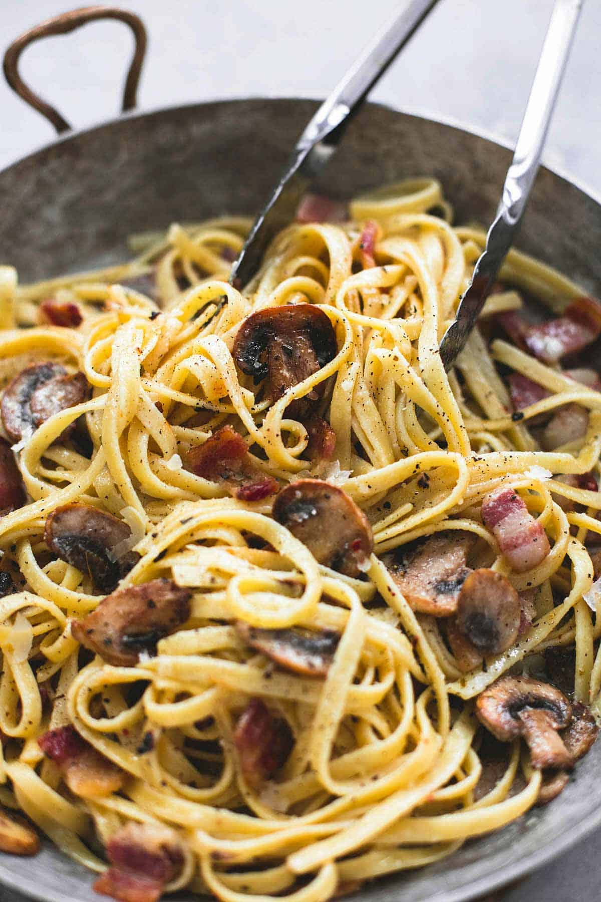 close up of parmesan bacon mushroom fettuccine with tongs in a wok.