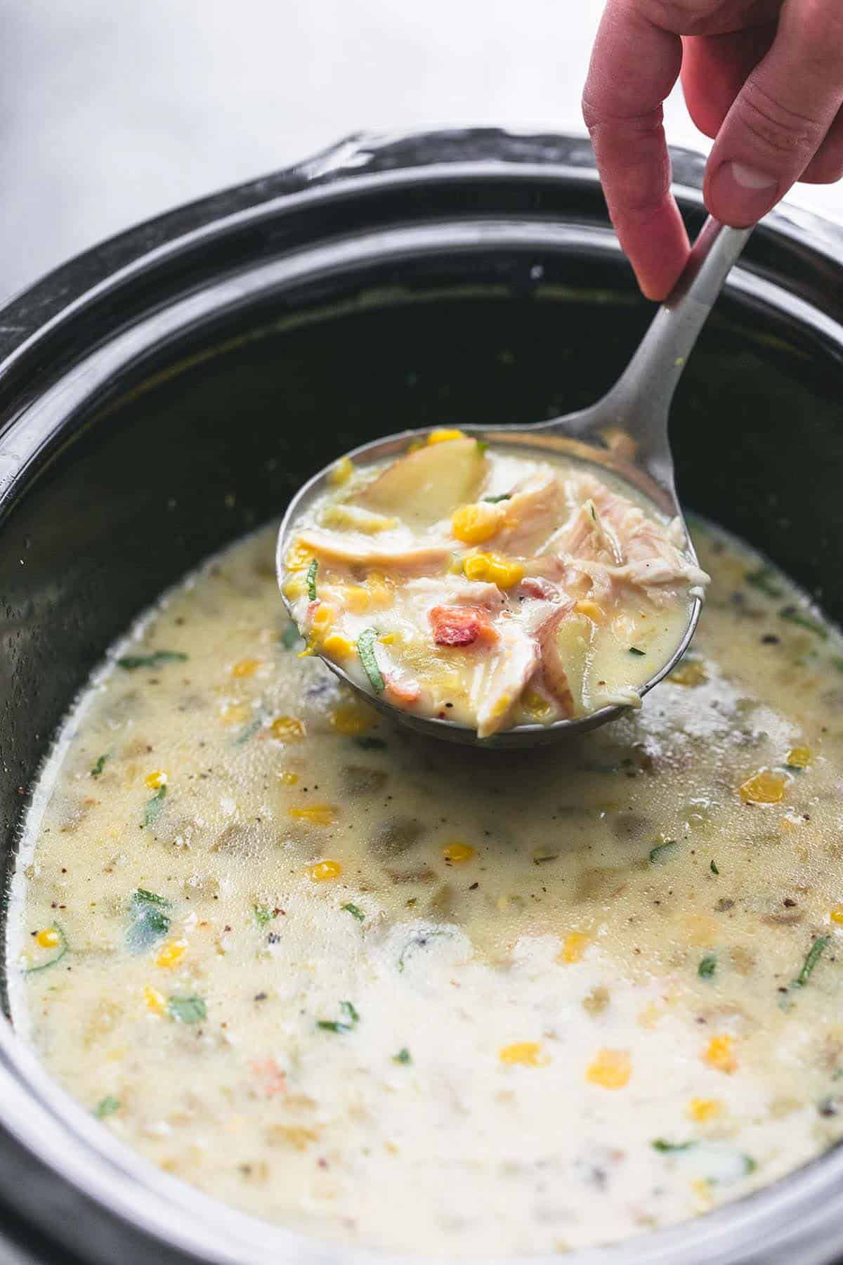 a hand holding up a serving spoon with slow cooker corn chowder in it above a slow cooker of more chowder.