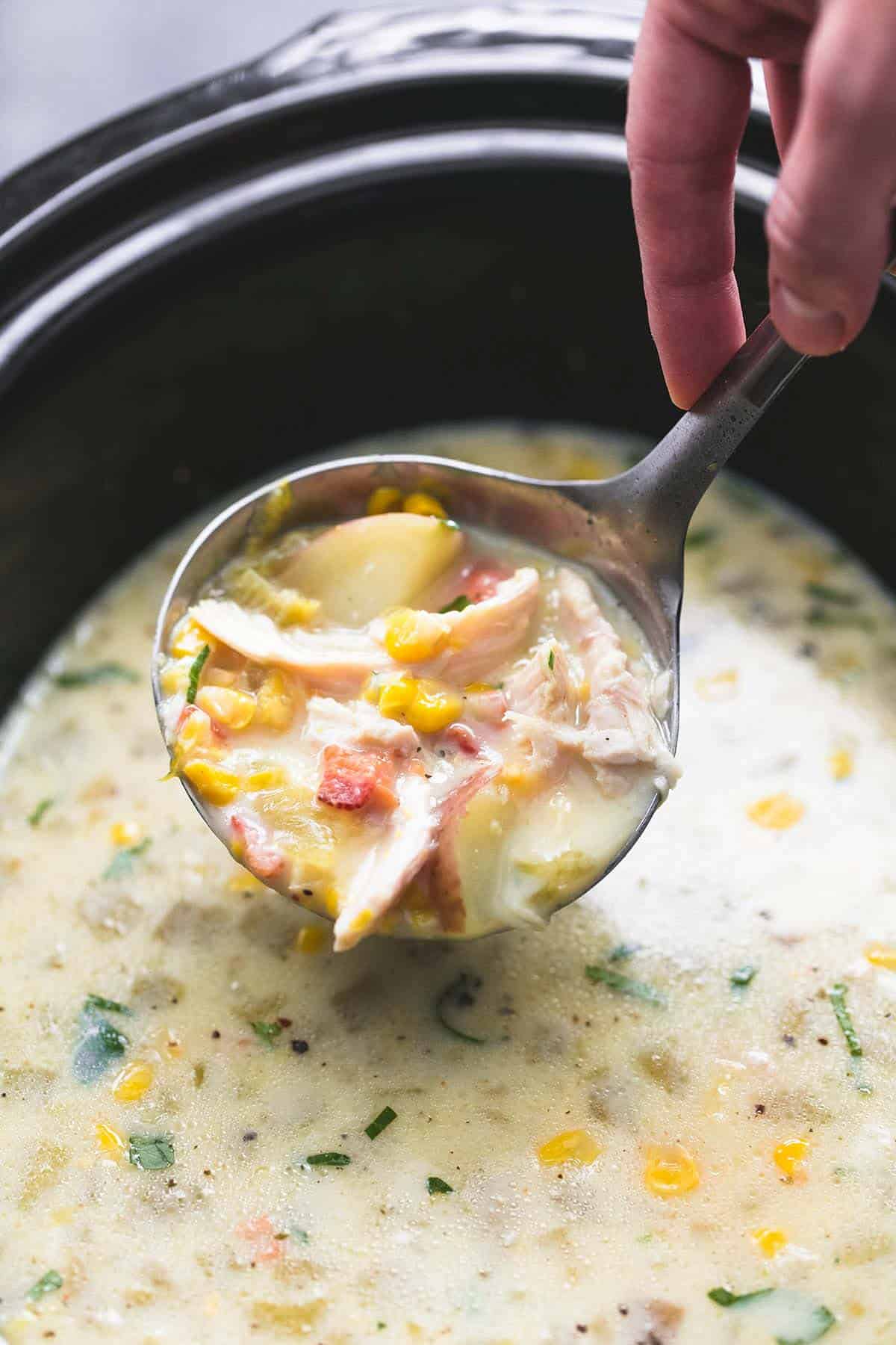 a hand tilting a serving spoon of slow cooker corn chowder above a slow cooker of more chowder.