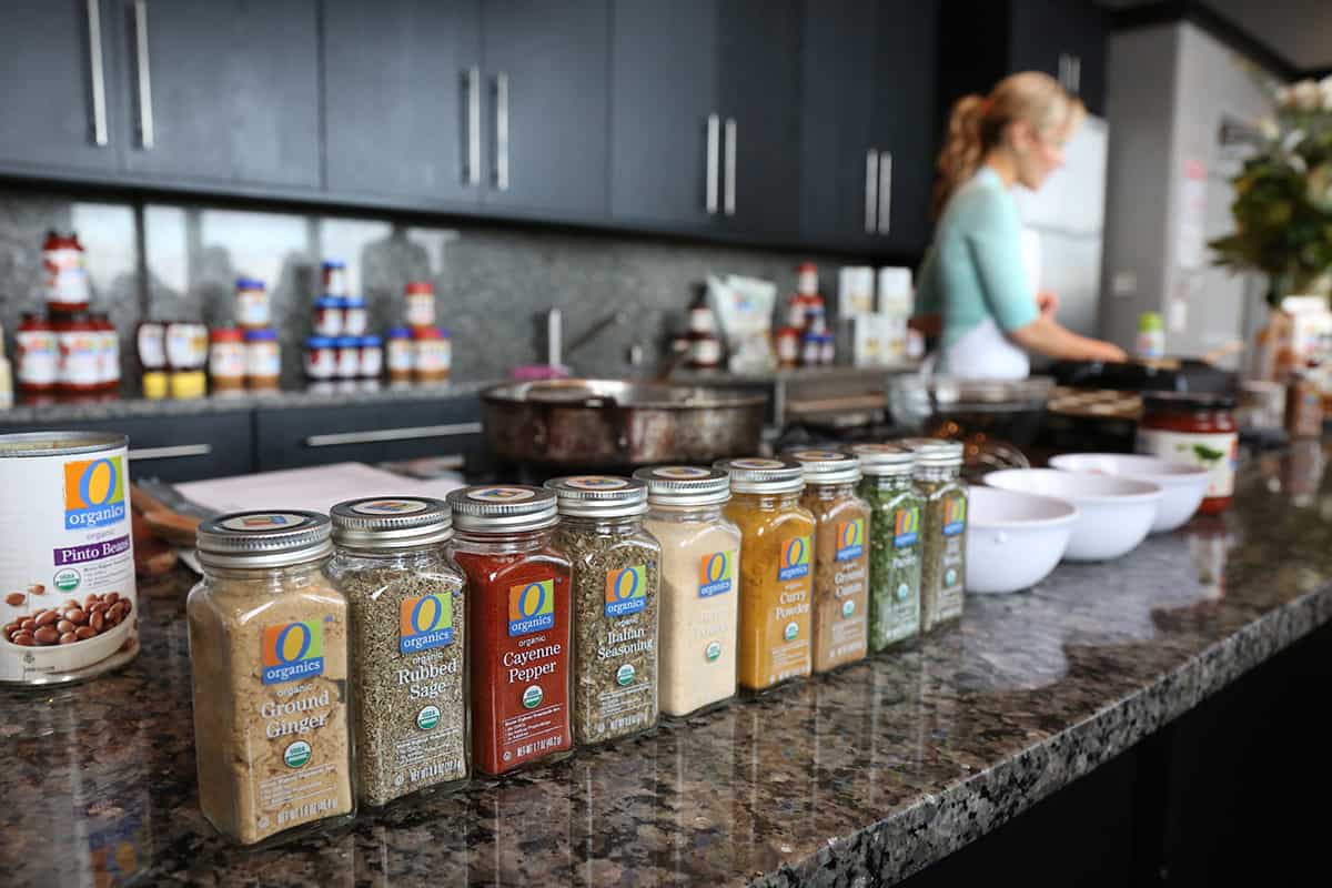 spice containers lined up on a counter in a kitchen with a women cooking off to the side.
