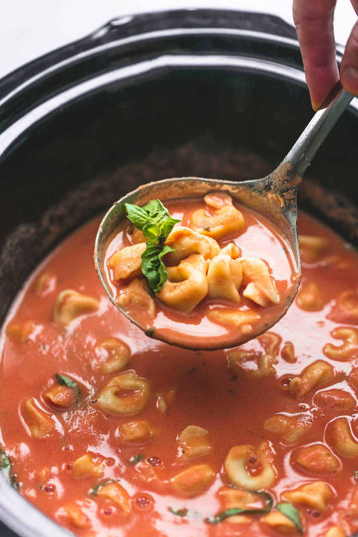a hand holding up a serving spoon of slow cooker tomato basil parmesan tortellini soup above a slow cooker with more soup.