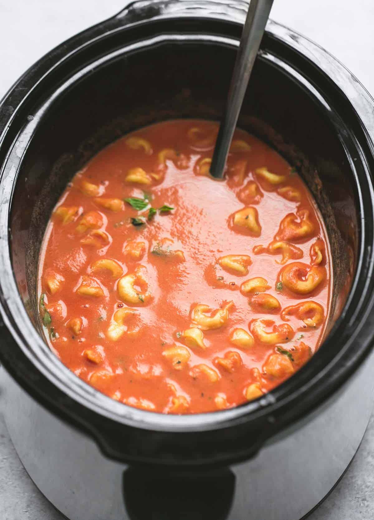 slow cooker tomato basil parmesan tortellini soup with a serving spoon in a slow cooker.