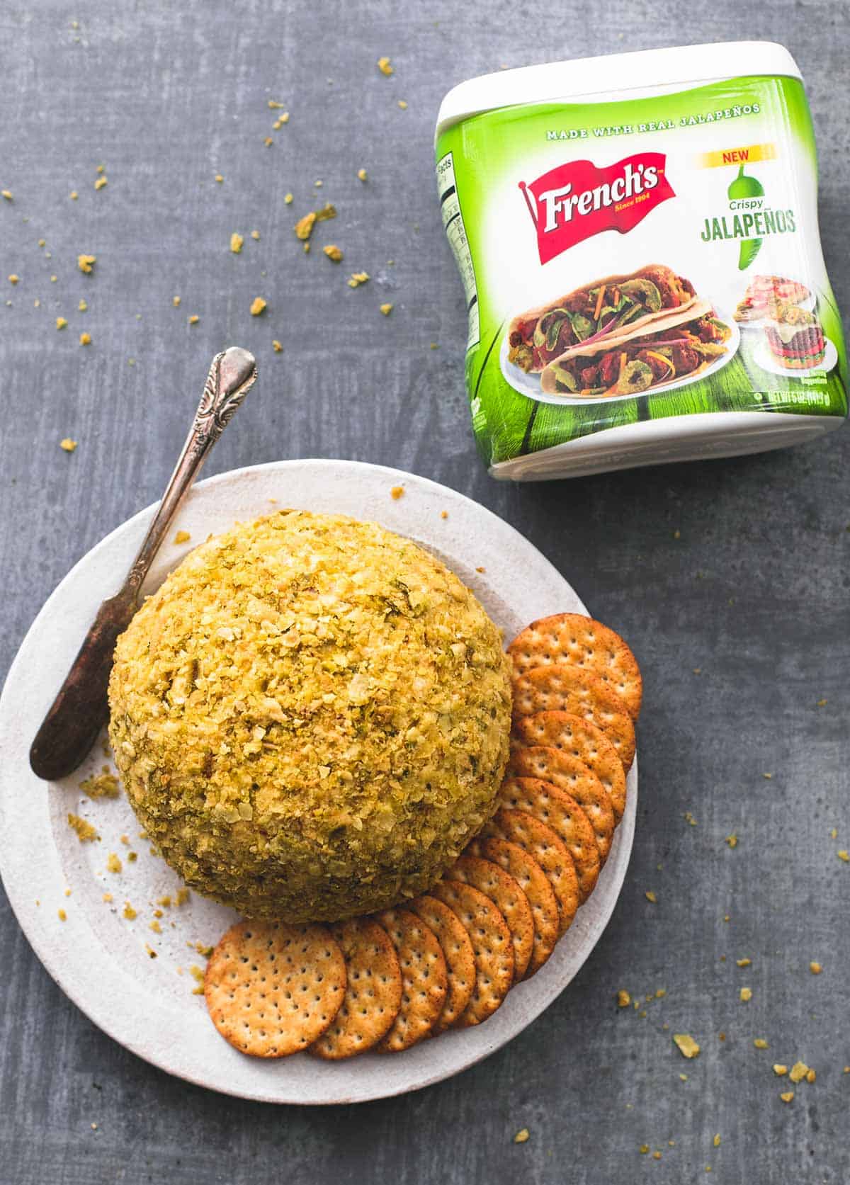 top view of cheddar bacon jalapeno cheeseball with crackers and a spreading knife on a plate with a container of dried jalapenos on the side.