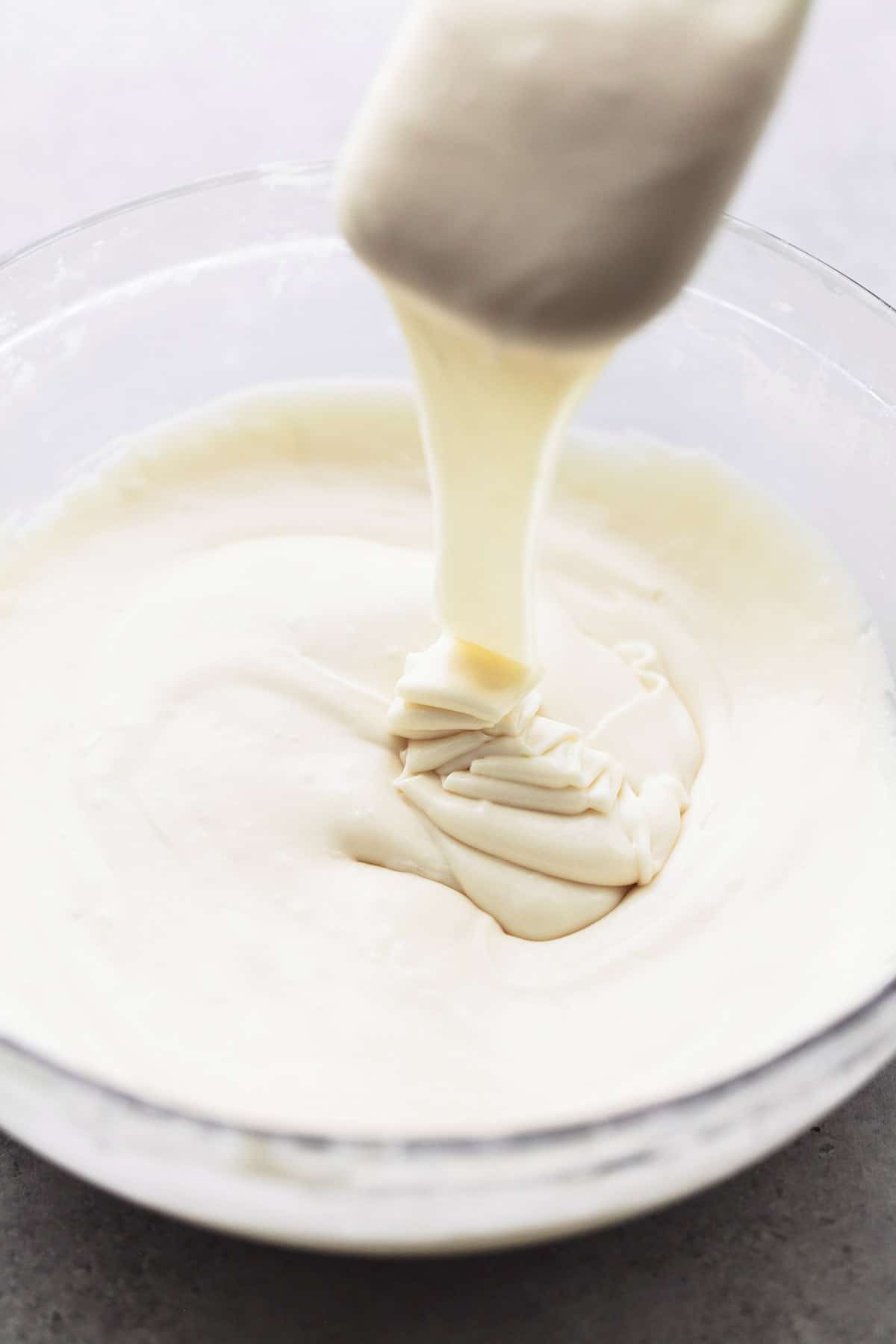 a rubber spatula being lifted from a glass bowl of cinnamon roll icing.