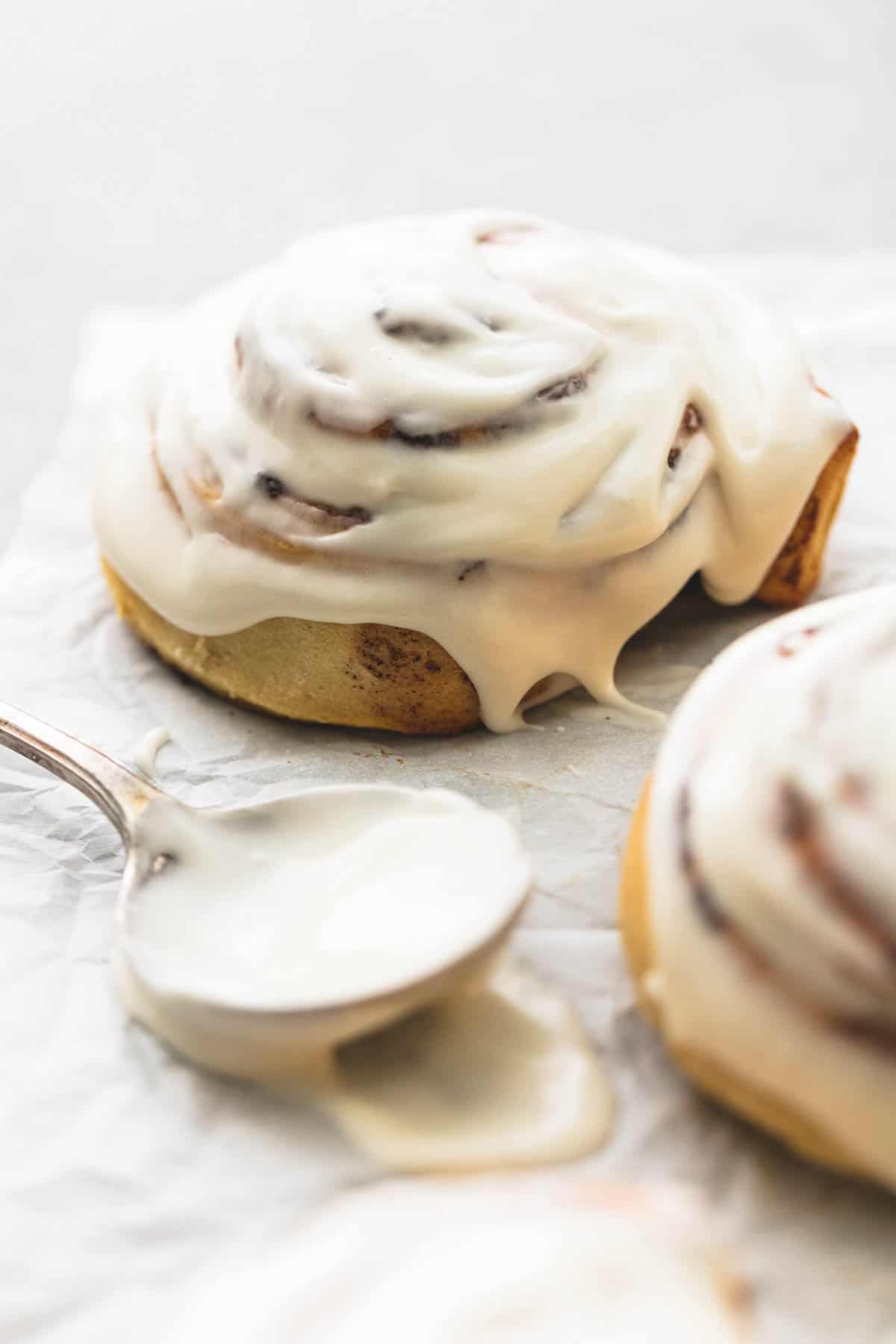 a spoon with cinnamon roll icing on it with an iced cinnamon roll behind it and on the side.