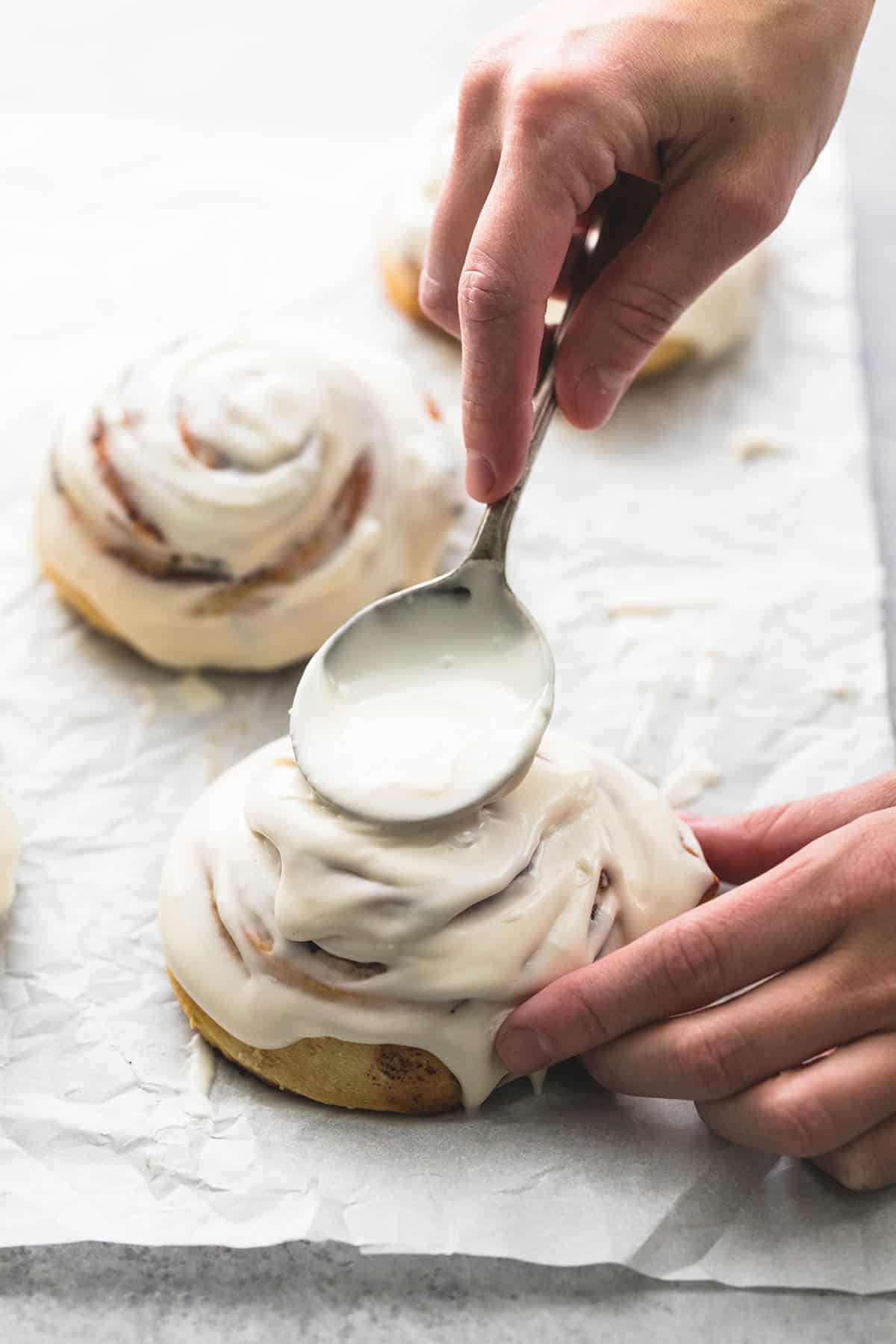 a hand icing a cinnamon roll with a spoon with more iced rolls in the background.