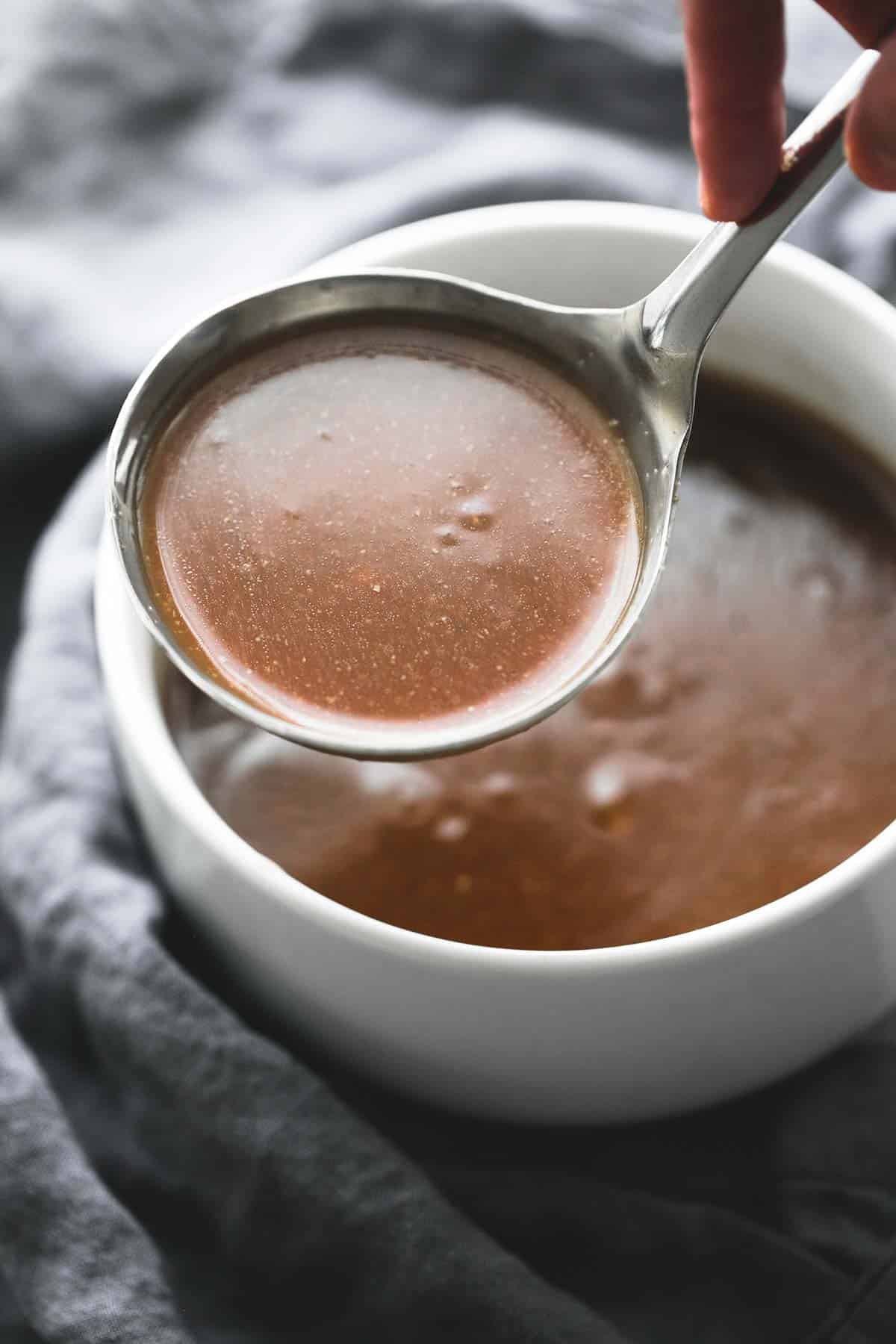 a hand holding up a serving spoon of beef broth brown gravy above a bowl of more gravy.